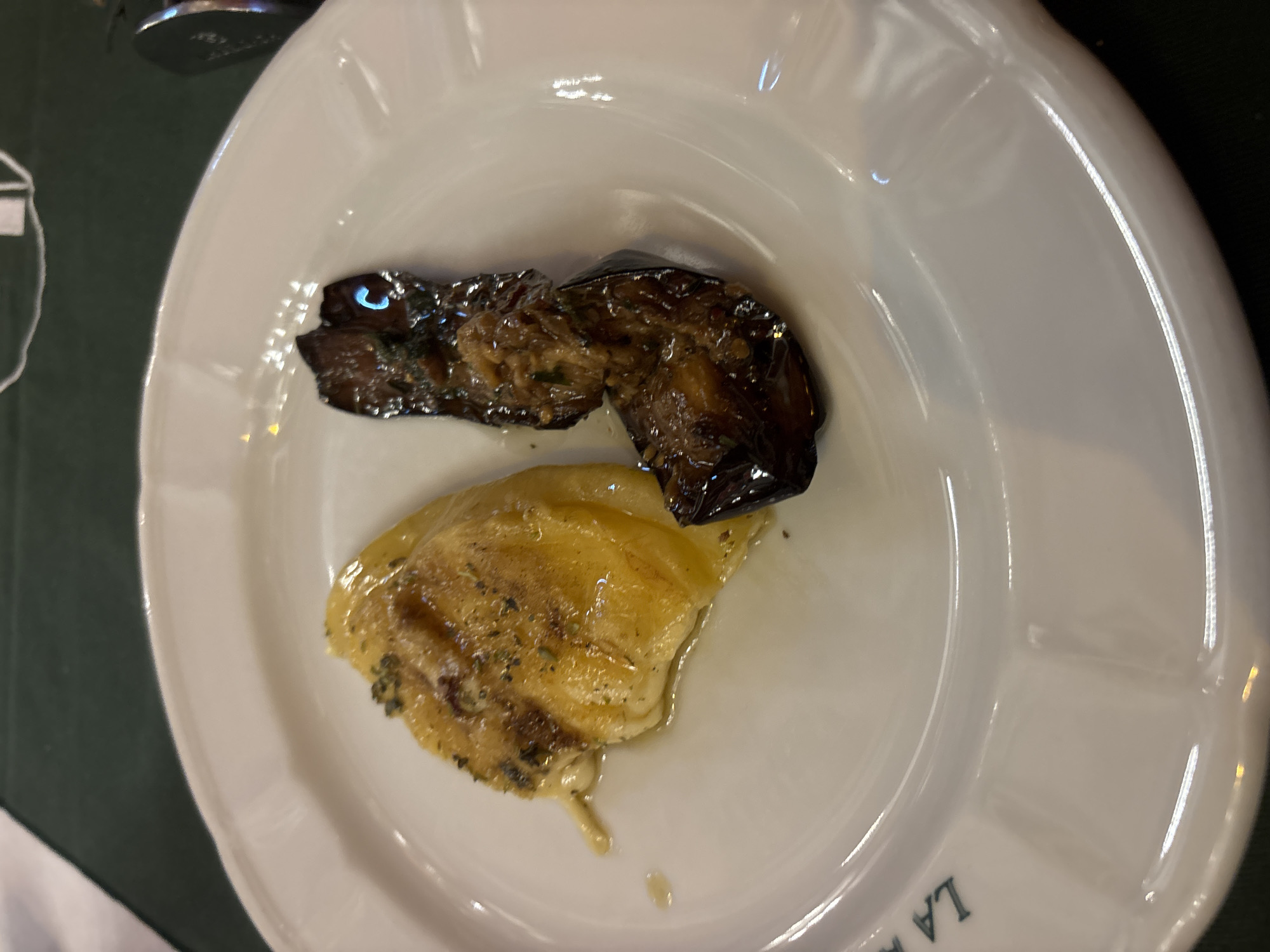 A white plate contains two food items. One is a dark eggplant, possibly grilled or roasted. The other is a lighter-colored, possibly cheesy or saucy, food item with herbs. The plate is on a dark green surface. The plate's edge reads "LA", suggesting it is at La Brigada Parrilla, a restaurant in the city of Buenos Aires, Argentina.