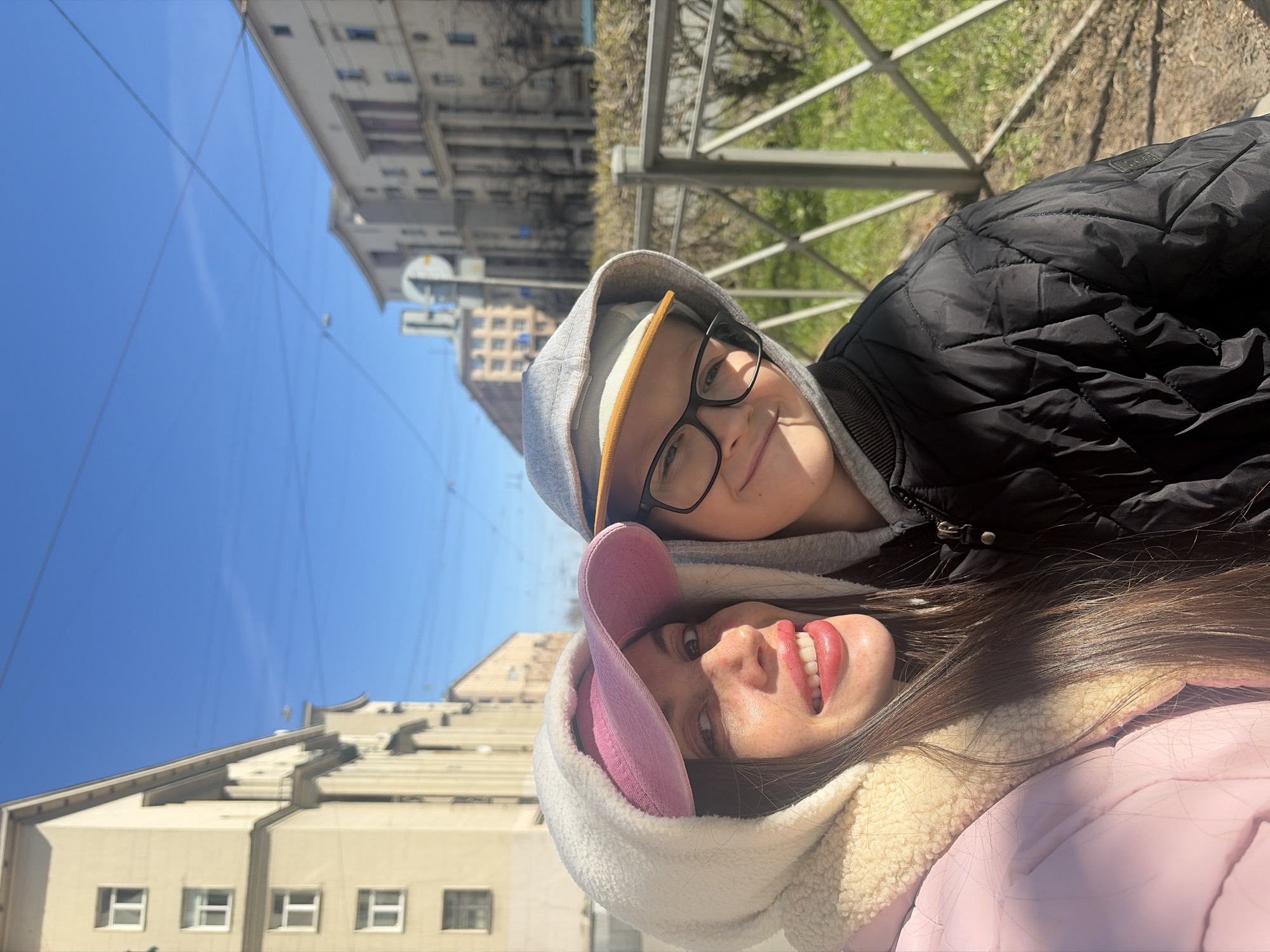 A selfie-style portrait, rotated 90 degrees clockwise, features two smiling individuals outdoors on a clear, sunny day. To the left, a woman with long dark hair is seen, wearing a white fluffy hooded jacket over a pink baseball cap and a light pink puffer jacket. She has red lipstick and a wide, joyful smile, looking directly at the camera. Next to her, a child or young person is visible, wearing a grey hooded sweatshirt over a light-colored baseball cap, black-rimmed glasses, and a black quilted jacket with an indistinct logo on the shoulder. They are also smiling and looking towards the camera.
The background, from top to bottom, shows a vibrant blue sky crossed by several thin overhead lines, possibly for trams or utilities. Below the sky, multi-story buildings with numerous windows and classic architectural details line what appears to be a street. Further to the right of the subjects, a metal railing is visible, with green grass and bare deciduous trees suggesting a park or public green space. The bright sunlight creates some shadows, particularly across the woman's face. Despite the sunshine, the subjects' hooded jackets and puffer coats indicate cool weather, likely early spring or late autumn. The urban setting and architecture are consistent with Saint Petersburg, Russia. Both individuals convey happiness and contentment.