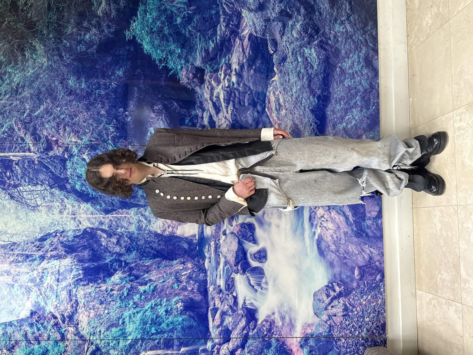A person is standing indoors in front of a large mural depicting a landscape with a blue-toned waterfall cascading down rocks. The individual is wearing a brown jacket with silver-toned buttons, a white collared shirt, and striped gray trousers. They are also wearing black shoes. The floor is tiled with a light-colored material. The overall scene appears to be a staged or posed photograph. The location is not discernible.