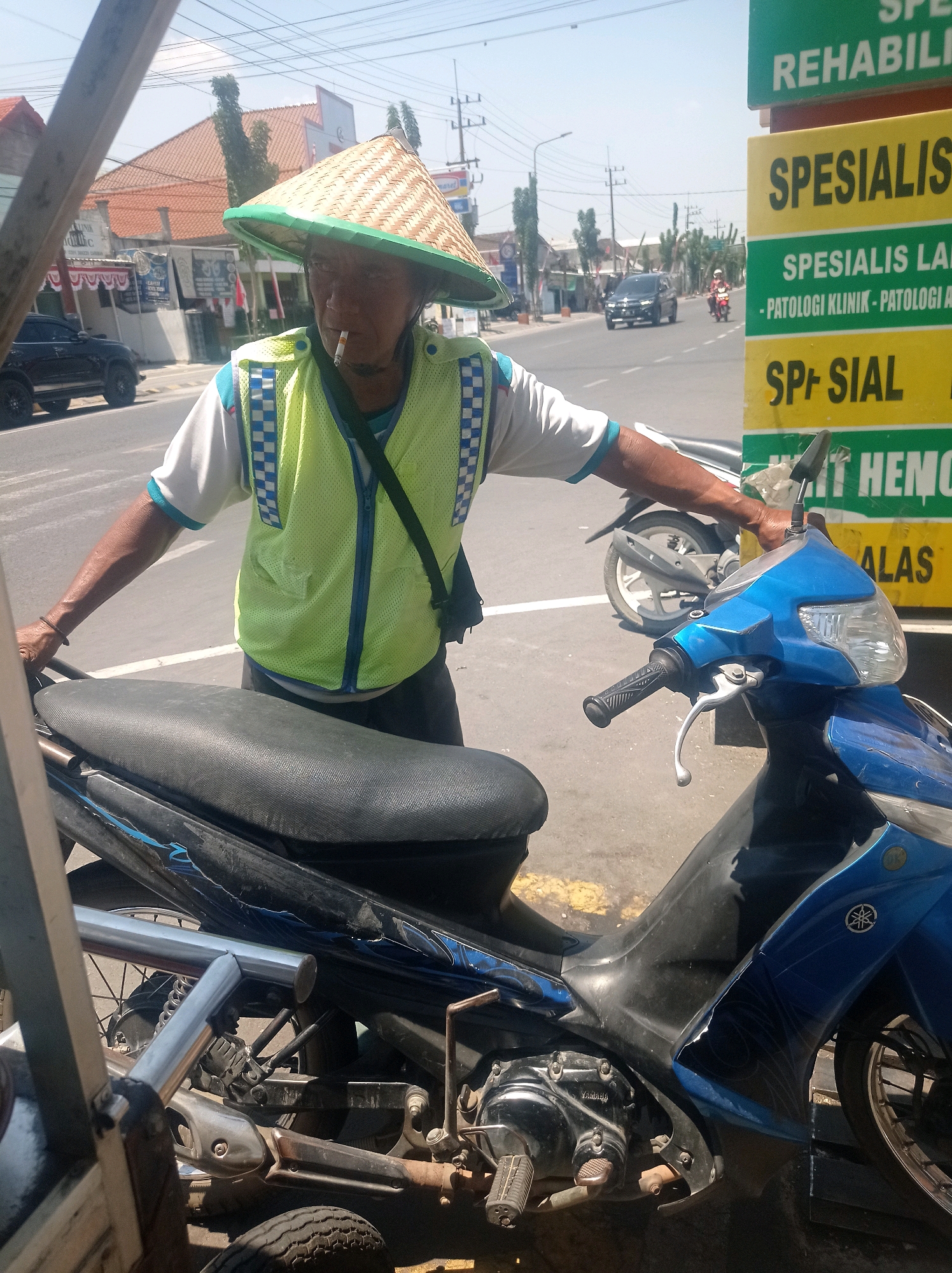 In Bangunsari, Indonesia, a man wearing a conical straw hat and a bright yellow reflective vest stands next to a blue motorcycle. He is smoking a cigarette and leaning on the motorcycle's seat. The man wears a white shirt with blue trim and a black strap diagonally across his vest. Behind him, traffic moves along a paved road under a clear sky. Signs with green and yellow lettering, some in Indonesian, are visible in the background. A black SUV is parked on the left side of the road, and other motorcycles are in motion. The scene appears to be a street-level view on a sunny day.