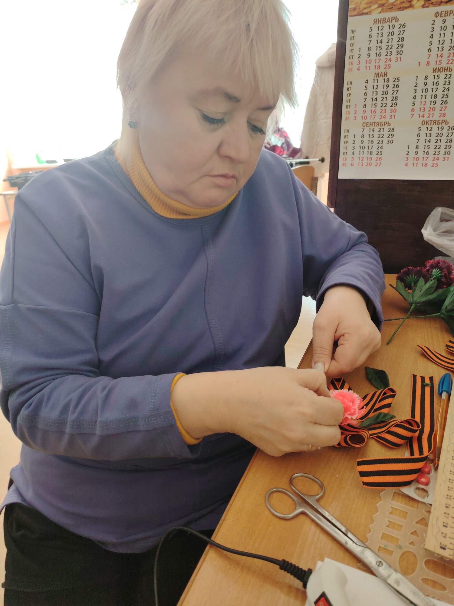 The image depicts a woman sitting at a table, crafting. She has short, blonde hair and is wearing a blue long-sleeved shirt over a yellow turtleneck. She is focused on her work, using her hands to assemble what appears to be a flower attached to a St. George ribbon.
On the table, there are scissors, additional St. George ribbons, flowers, a stencil, a pen, and a glue gun (plugged in). A calendar is hanging on the wall behind her; the months of January, May, September, and October are visible.
The information that the image was taken in Sarapul, Russia is not visually discernable.