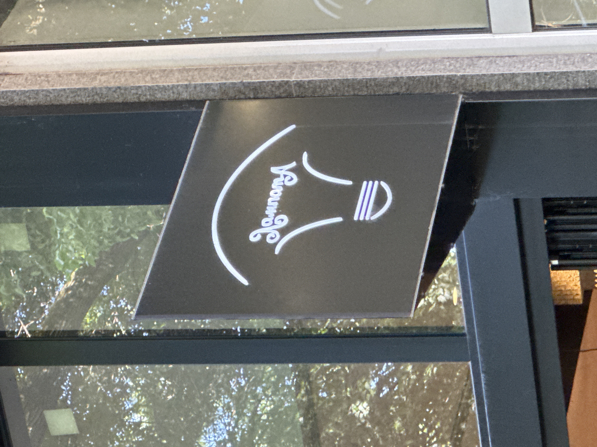 A close-up shot shows a black sign with a white logo resembling a lightbulb and the name "Somara" written vertically. The sign is attached to a dark gray metal frame, with a window and greenery visible in the background. The image was taken in Sofia, Bulgaria.