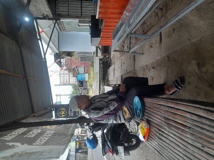 The scene depicts an outdoor roadside area in the city of Pandanarum, Indonesia, under a corrugated metal roof supported by wooden and metal beams. A single adult male is seated on a long, slatted wooden bench on the left side of the frame, facing right and slightly upwards. He wears a light-colored skullcap, a purple and grey long-sleeved jacket, black trousers, and black and white striped slide sandals. A dark scooter with a blue helmet on its seat and license plate "W 2266 WF 01.29" is parked behind him. On the bench next to him are a multi-colored tissue box and a light blue plastic bowl containing dark green material. The ground is dirt. To the right, a long, low metal bench or table is present. Behind it are tall orange structures. The background reveals a paved road with a white line, various buildings including a grey-walled structure, a red-bricked building, and a light-colored building with red and white striped ornamentation, utility poles, and distant vegetation under an overcast sky. A dark banner with light text and a circular "YKS" logo hangs from the roof structure on the left, and a bright light fixture is affixed to the roof on the right.