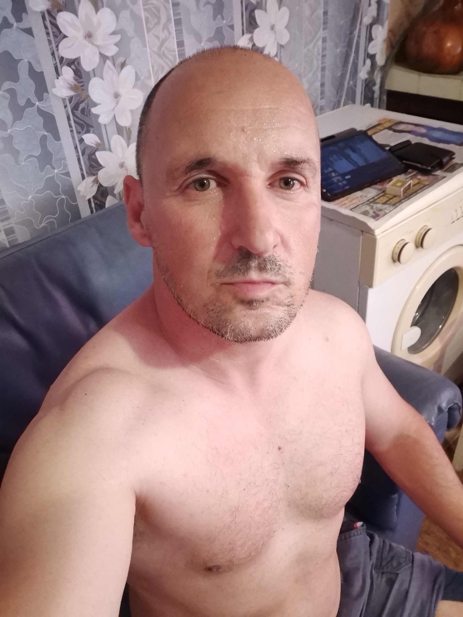 A man with a bald head, dark stubble beard, and light-colored eyes (possibly green or hazel) is captured in a selfie-style shot. He is shirtless, revealing a broad chest and muscular shoulders, with his skin appearing slightly shiny, possibly from sweat. He wears dark shorts or pants, visible around his lap. His expression is neutral as he looks directly at the camera.
The man is seated on a blue, possibly leather, sofa or armchair. In the background to his left, there is a curtain featuring a pattern of large white flowers, likely magnolias, on a lighter, possibly striped or patterned fabric. To his right, a white washing machine is visible, with a tablet device, a dark wallet, and a small white object placed on a surface above it, possibly a magazine or newspaper. The tablet screen displays some illuminated content. The setting appears to be an indoor, domestic environment, potentially a living area or a utility space given the presence of the washing machine. No discernible text is visible in the image.