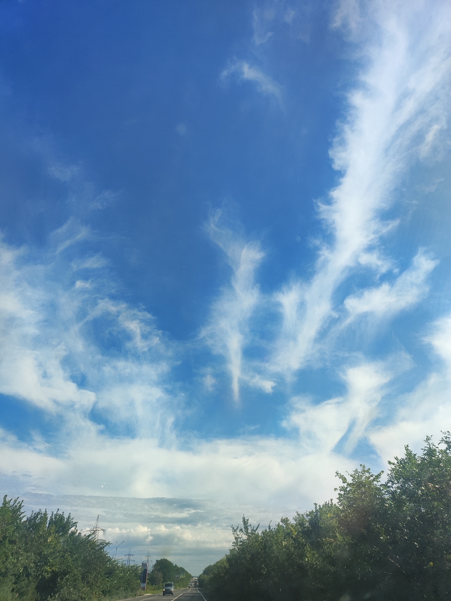 The image captures a clear blue sky filled with wispy white clouds, seen from the perspective of a moving vehicle on a road. The sky occupies the majority of the frame, with the clouds forming intricate patterns. Below the sky, a strip of green trees lines both sides of the road, which is asphalt and appears to be a highway. In the distance, there are a few vehicles on the road, and power lines and transmission towers are visible behind the trees on the left. The overall scene suggests a rural or suburban setting during daylight hours.