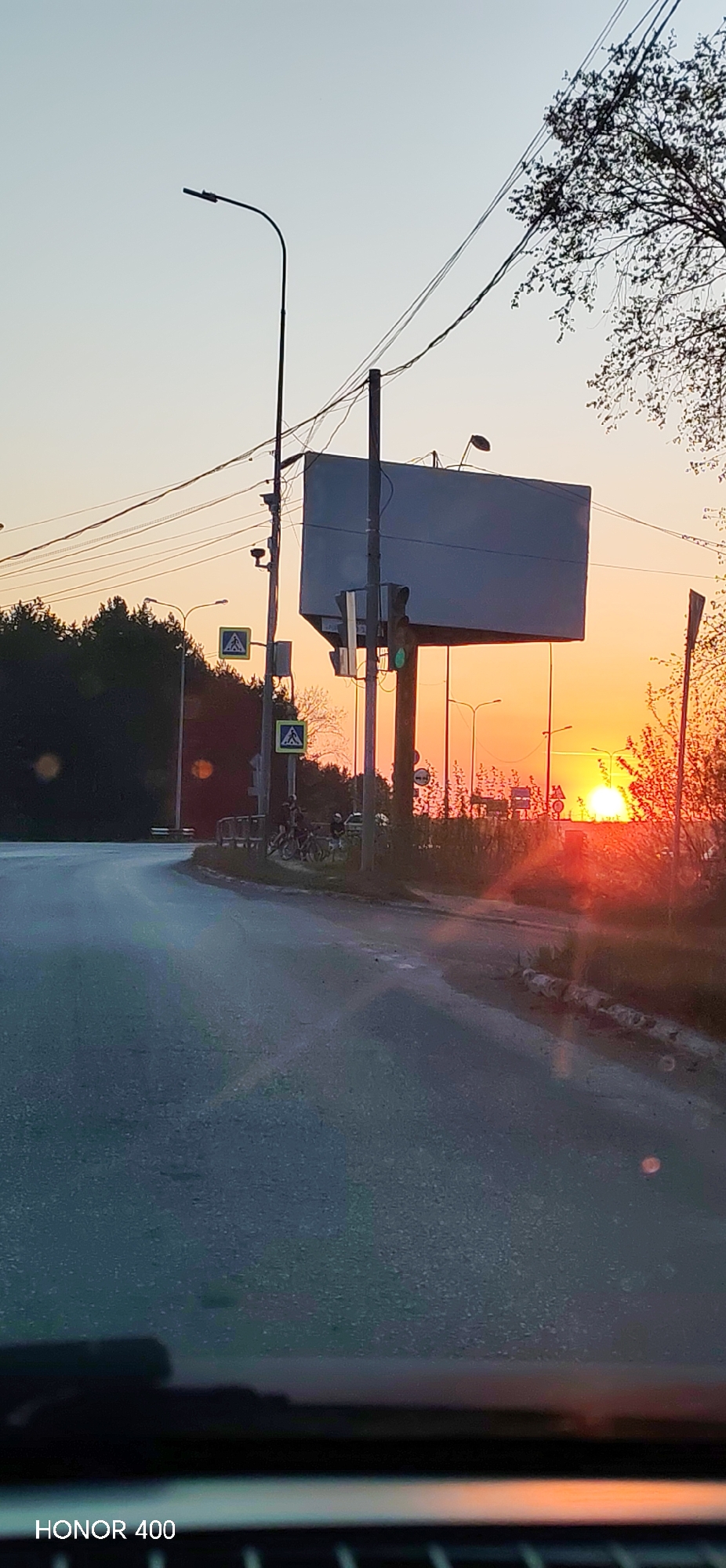 The photograph was taken on a road in the city of Chaikovsky, Russia, at sunset. The sun is situated low on the horizon, creating an orange glow across the sky. A large, rectangular billboard is positioned above the road, supported by a central pole. A traffic light displaying green is mounted to the side of the billboard. In the vicinity of the billboard, there are street lights, power lines, and pedestrian crossing signs. Some individuals with bicycles are gathered near a barrier to the side of the road, next to the pedestrian crossing. The road itself curves gently. The image was taken from inside a vehicle, as indicated by the dashboard visible at the bottom.