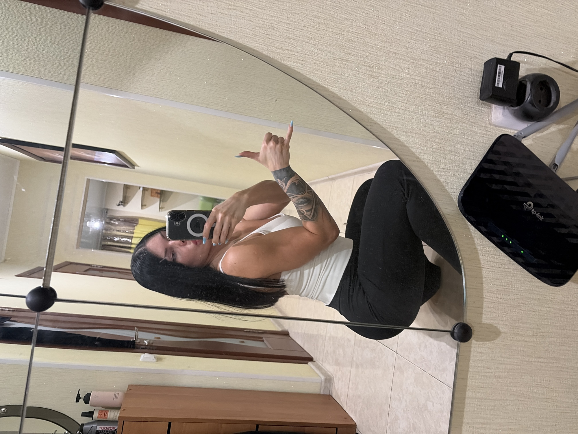In a domestic setting, a woman with long, dark hair, wearing a white tank top and black leggings, poses for a selfie in a mirror. She is in a crouched position, holding a smartphone in her right hand, and her left hand is making a "shaka" gesture. Her left arm is covered in tattoos. The background of the reflection shows a hallway with yellow walls, a wooden door, and shelves. In the foreground of the original image, a black TP-Link router and a small black electrical device plugged into a wall socket are visible on the floor. The floor is tiled. There is no visible indication of the location being in Ufa, Russia.