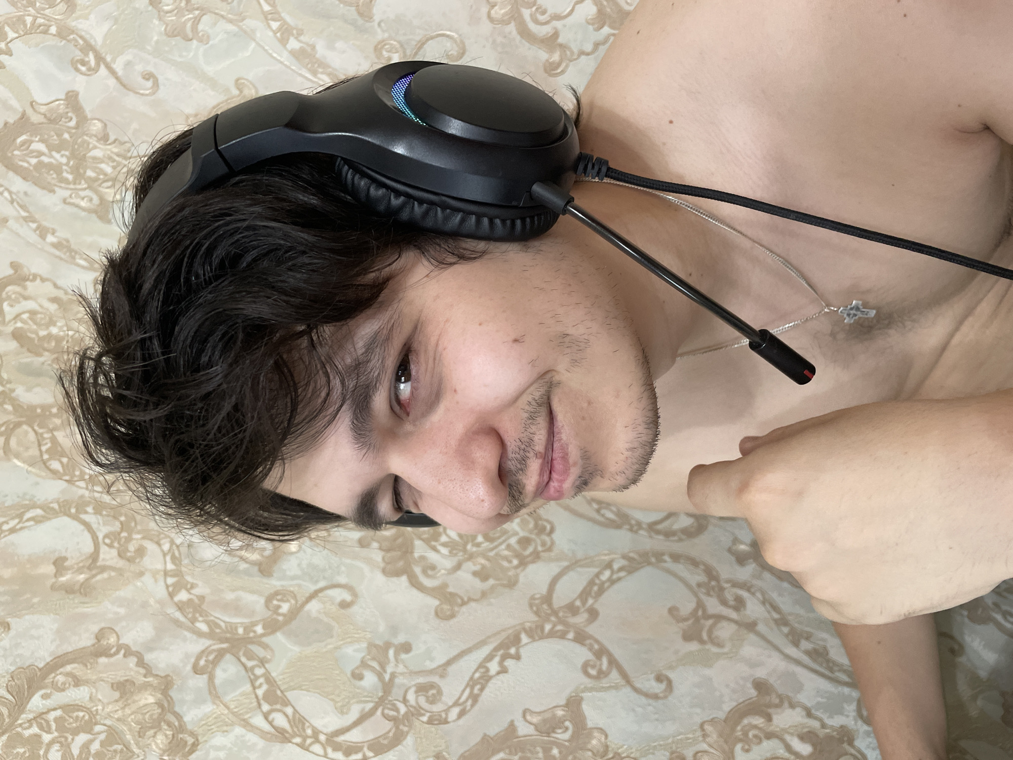 A man lies on a bed, wearing black headphones. He has dark hair and light skin. A silver necklace with a cross pendant rests on his bare chest. His right hand is visible near his torso. The bed is covered with a patterned beige and white sheet. The image does not provide enough information to determine the location in Izhevsk, Russia.