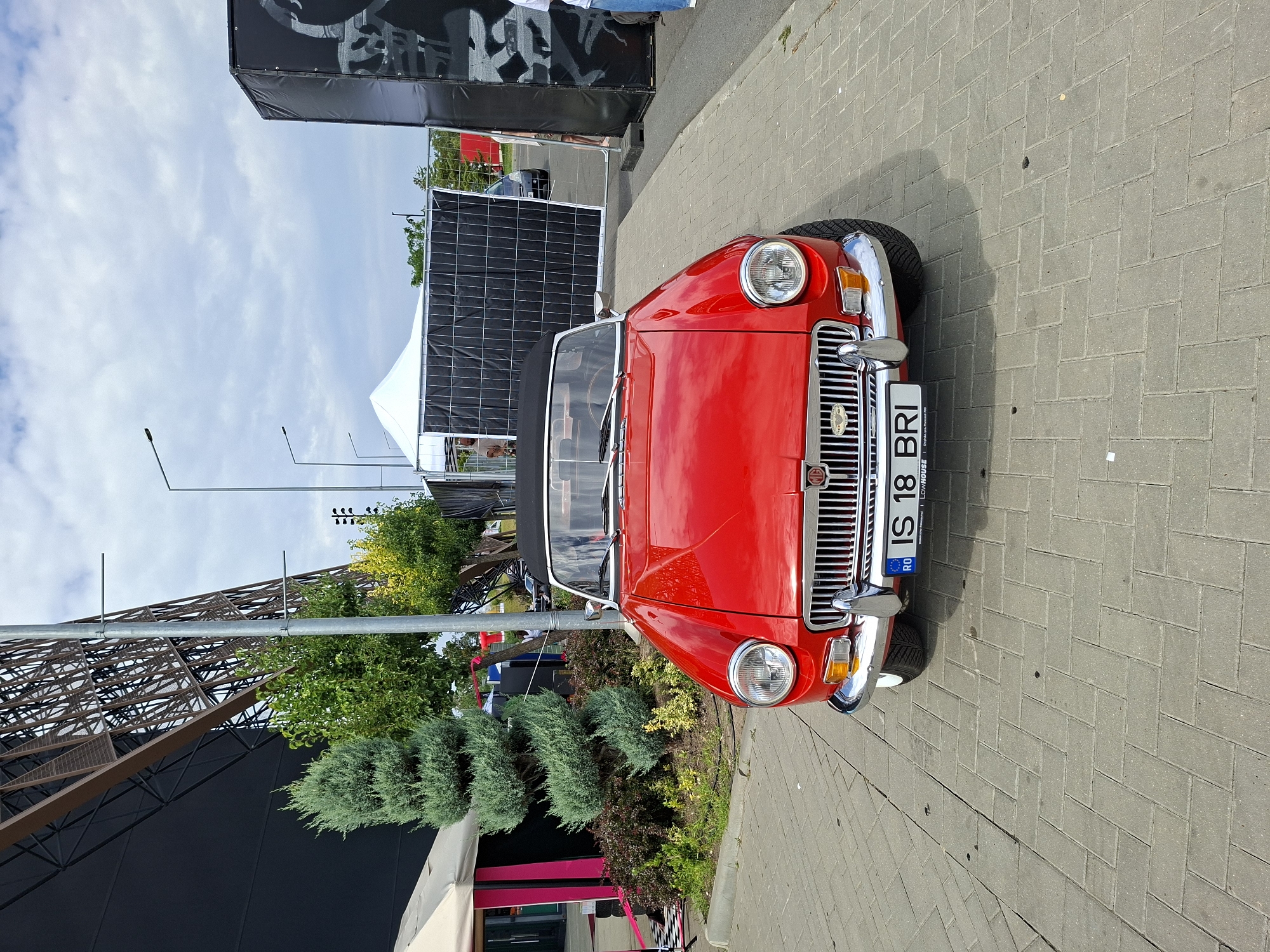 An outdoor, eye-level, medium shot captures the front of a bright red, vintage convertible MG car parked on a brick pathway. The car is the primary subject, with its chrome grille, round headlights, and license plate clearly visible. The license plate reads "RO IS 18 BRI". Behind the car, a large, dark structure with a geometric pattern dominates the background on the right. To the left of the car, a large, black, tent-like structure with pink accents is partially visible. Several trees, including a conical evergreen, are planted between the car and the structures. The sky above is cloudy. The setting appears to be a public space in the city of Stăuceni, Moldova.