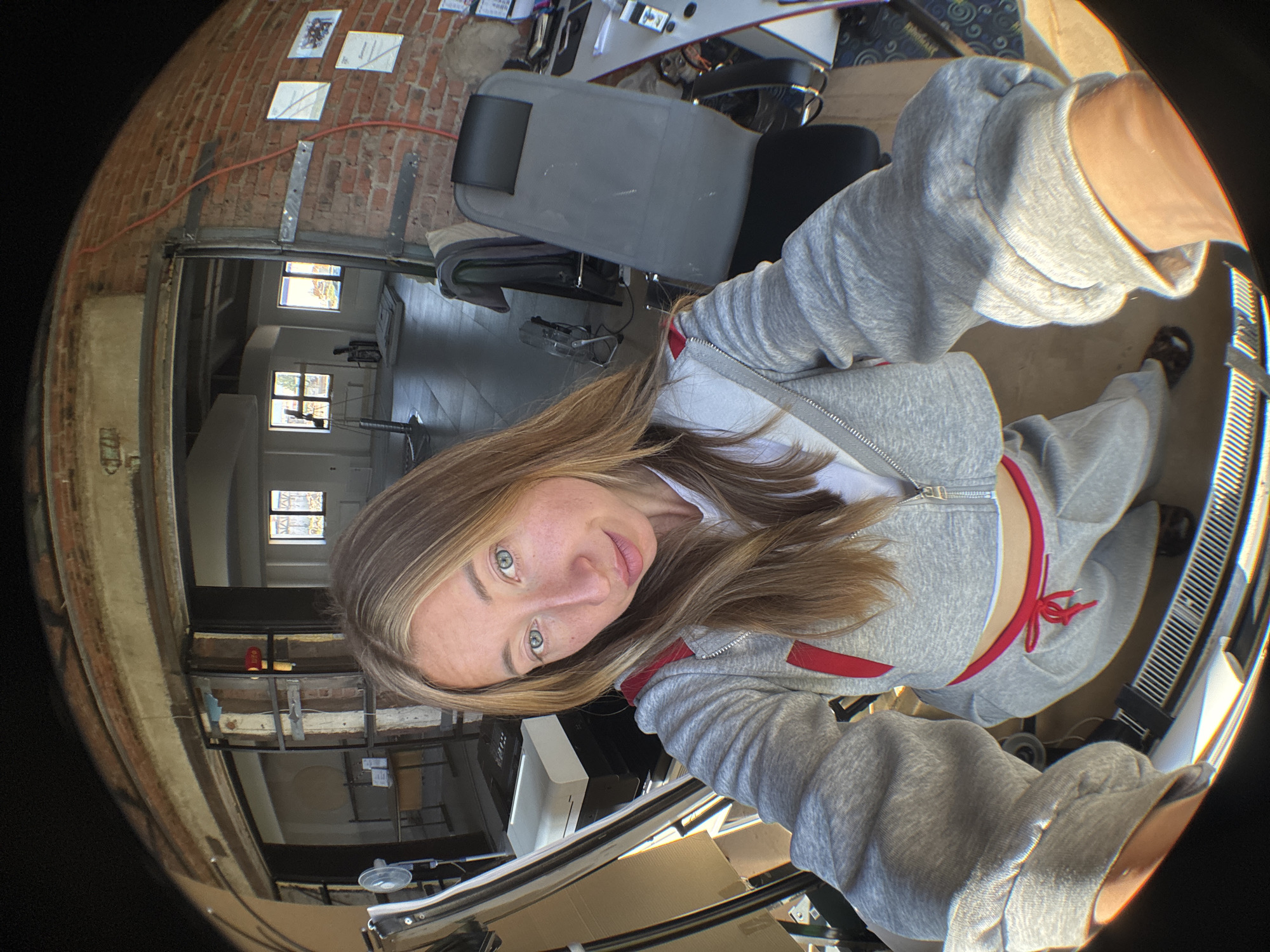 A young woman with blonde hair and blue eyes, wearing a grey hooded tracksuit with red accents, takes a selfie in what appears to be a construction or renovation site. The background shows exposed brick walls, wooden beams, and open doorways leading to rooms with windows and some office furniture. The image is captured with a fisheye lens, distorting the perspective and creating a wide-angle view of the environment. There are stacks of boxes and various construction materials visible, suggesting ongoing work. The woman's attire is casual, and her expression is neutral, looking directly at the camera. The location is specified as Chaikovsky, Russia.