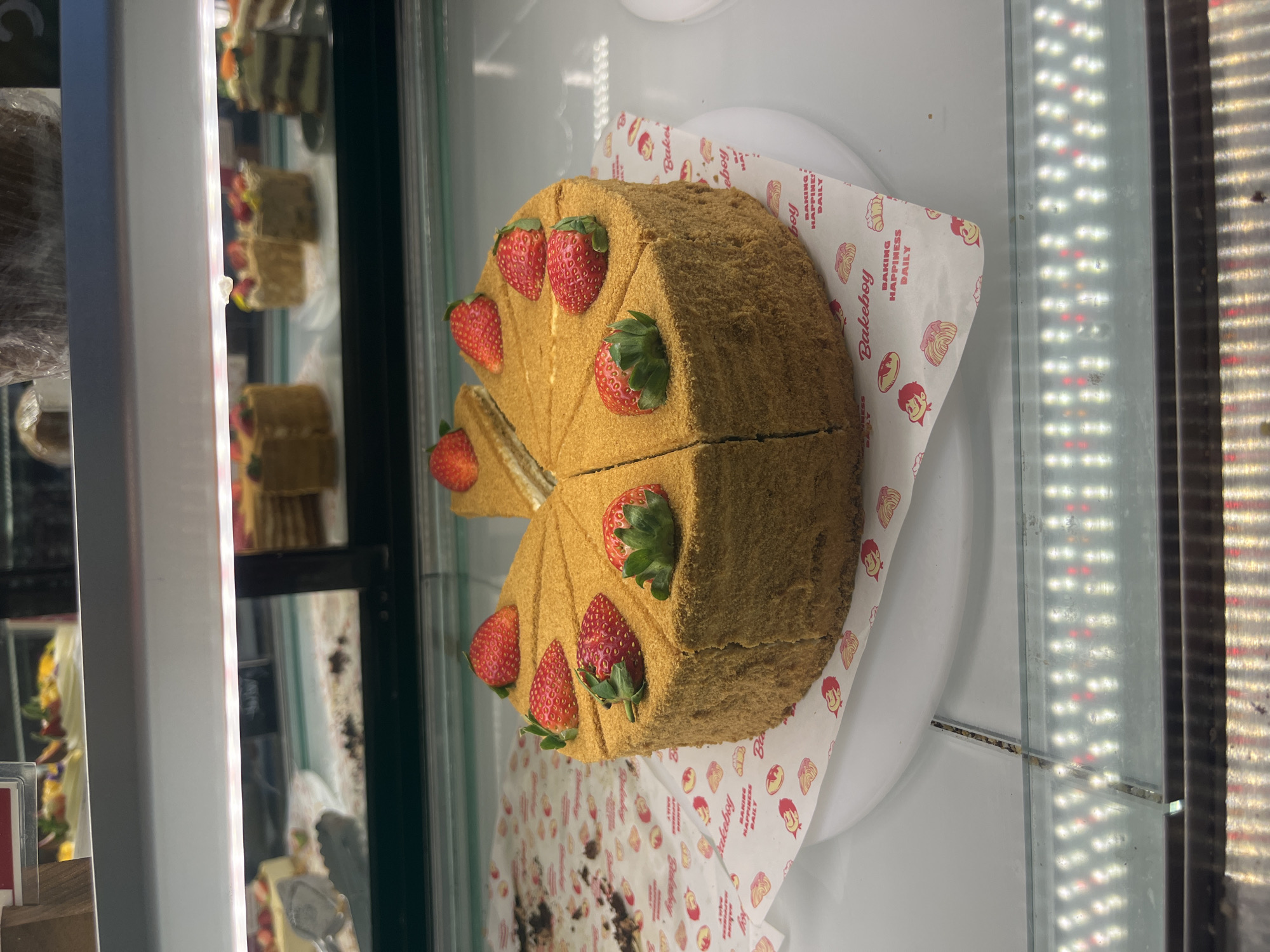 A honey cake with strawberries sits on a plate in a display case. The cake is cut into slices, with one slice removed. The plate rests on a piece of branded paper with the name "Bakeboy" and other designs. The display case is brightly lit with vertical LED strips. Other cakes are visible in the case. The city and specific location are unknown.