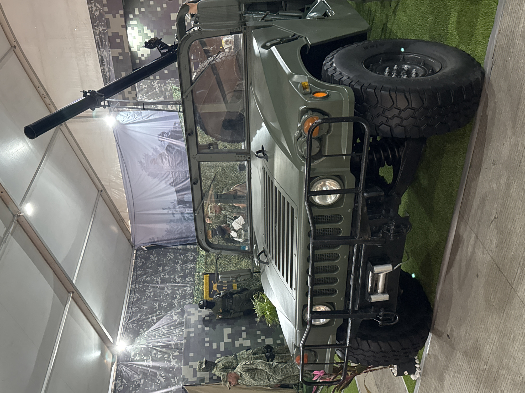The image shows a Humvee military vehicle, olive green in color, with a large black gun mounted on top. The vehicle is on display, possibly in a showroom or exhibition space. In the background, there's a camouflage pattern that could be a backdrop or part of the exhibition setup. There are also glimpses of people, at least one in a military uniform, looking at or near the vehicle. The location appears to be indoors, possibly in Puebla, Mexico, given context clues not explicitly visible in the image.
