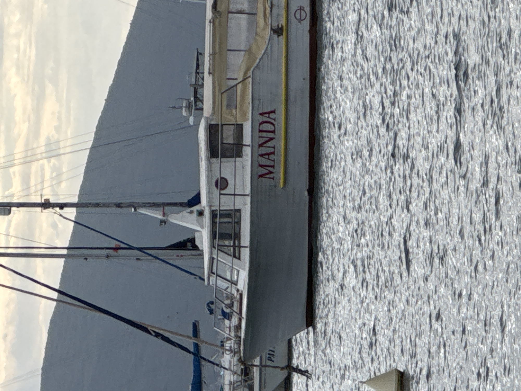 A slightly rotated, medium shot captures a boat named "MANDA" docked in Tivat, Montenegro. The boat is painted light gray with "MANDA" in red lettering on its side. The water's surface shows subtle ripples and reflects the light. Behind the boat, a mountain is visible against a sky filled with soft clouds. Mooring equipment is at the right edge of the shot.
