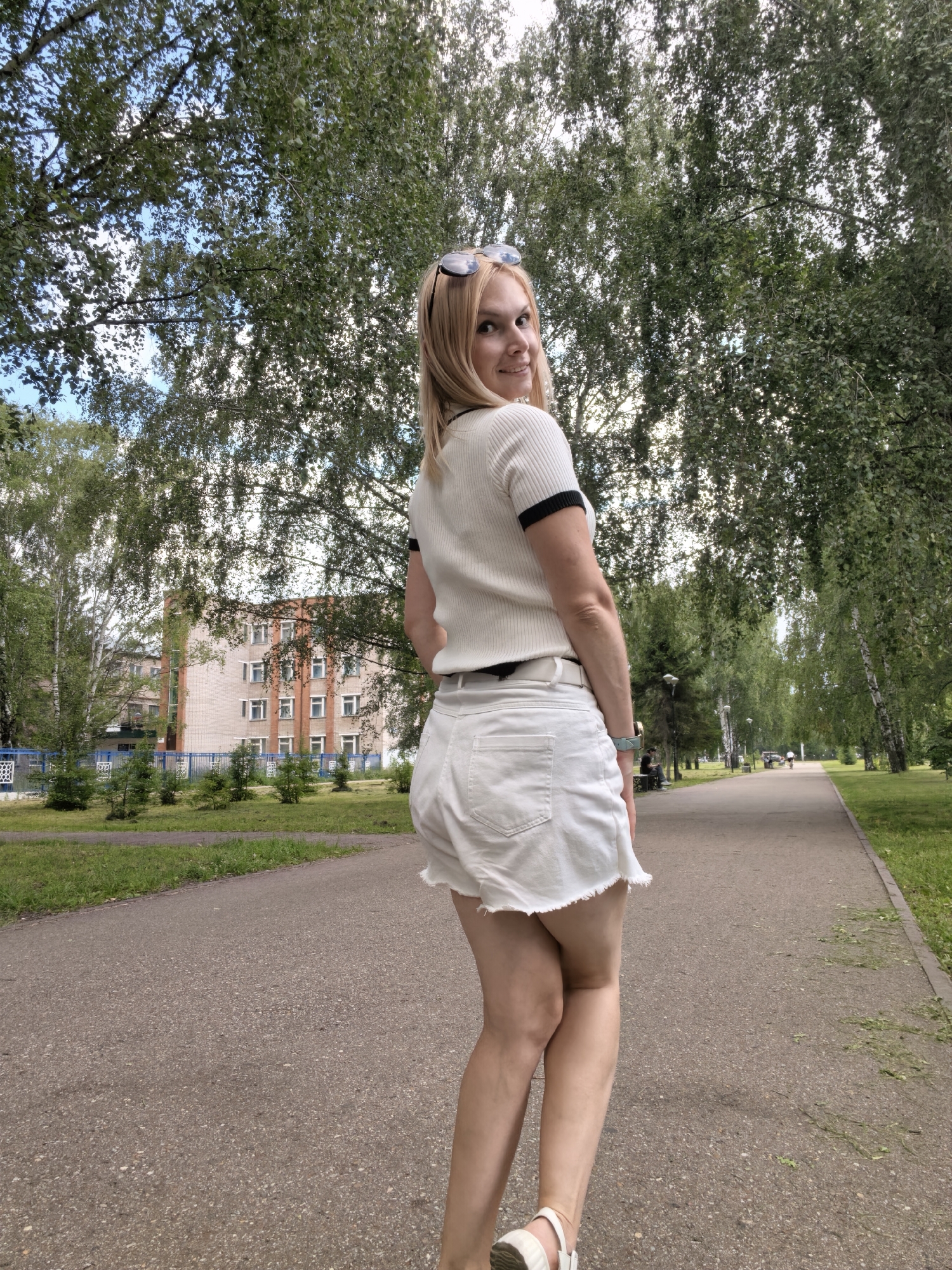 A blonde woman with sunglasses perched on her head is posing for a photo in a park. She is wearing a white short-sleeved top with black trim on the collar and sleeves, and white denim shorts. She is standing on a paved pathway with a grassy area to her left. In the background, a multi-story brick building with numerous windows is visible behind a blue metal fence. Large trees with green foliage line the pathway. The sky is partly cloudy. The location is the park on Oktyabrya Avenue in Sterlitamak, Russia.