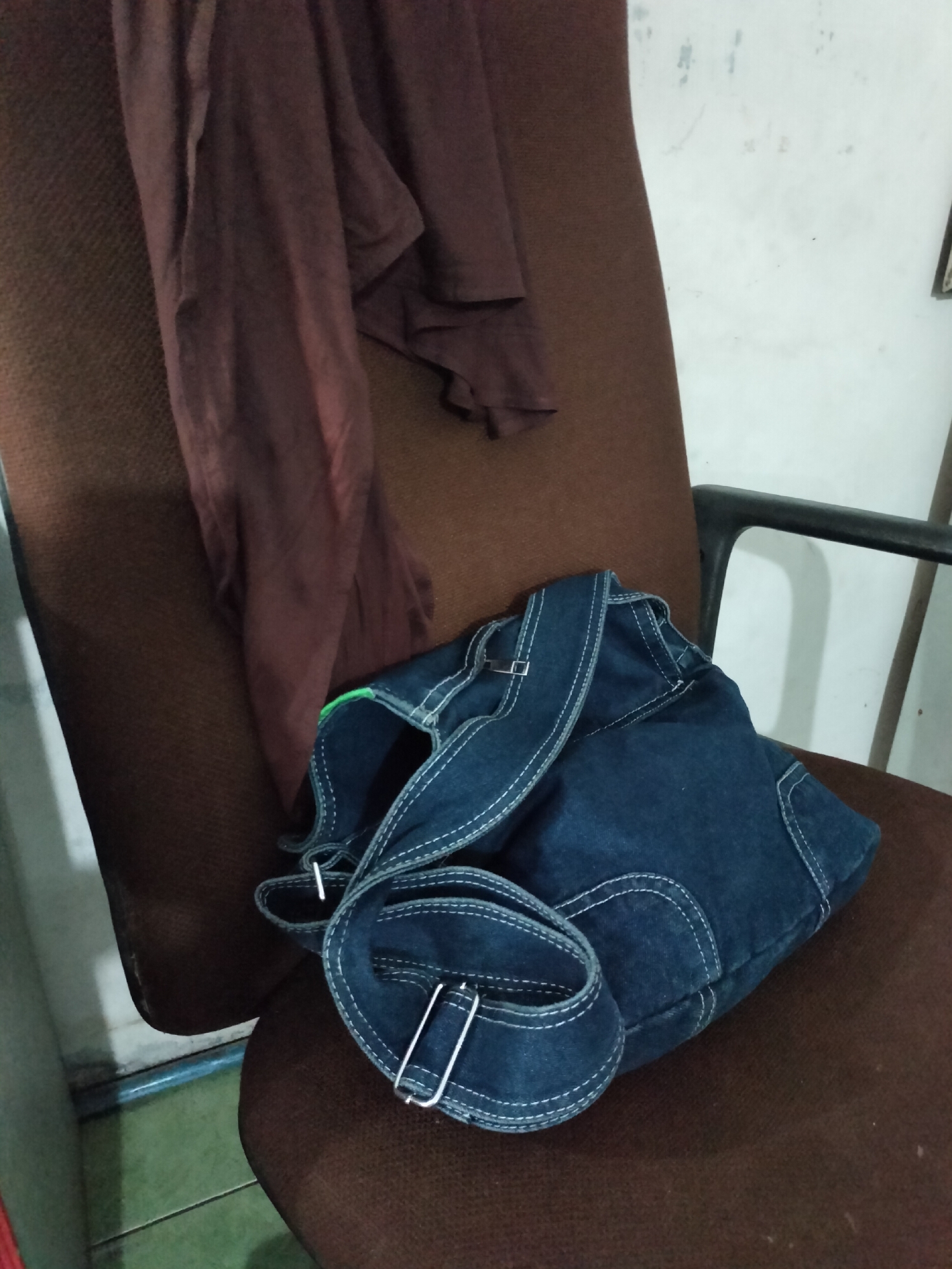 A dark brown office chair is seen from a slightly elevated angle. A dark brown garment is draped over the back and seat of the chair on the left side. In the foreground, on the seat of the chair, is a folded pair of dark blue denim overalls. The overalls have white contrast stitching and metal clasps on the straps. The background shows a white wall with some scuff marks. The floor is partially visible at the bottom left, with a green surface and a thin blue line. This scene is situated in an indoor location, possibly a home or office, in Langsa, Indonesia.