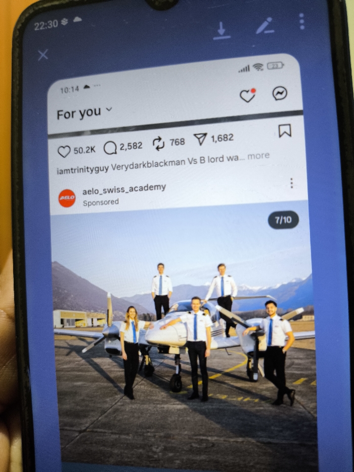 The image displays a close-up photograph of a smartphone screen showing an Instagram post. The smartphone, primarily black with a visible notch at the top, has a yellow object partially visible along its left edge. The phone's status bar shows the time "22:30," along with Wi-Fi, cellular signal, and battery indicators.
The Instagram interface includes a 'For you' header, a timestamp "10:14," and engagement metrics: 50.2K likes, 2,582 comments, 768 shares, and 1,682 saves. The post is from "aelo_swiss_academy" and is marked as "Sponsored." A user handle "iamtrinityguy" and truncated text "Verydarkblackman Vs B lord wa..." are also visible above the sponsored post. A "7/10" indicator in the bottom right corner suggests this is the seventh image in a carousel post.
The central image within the Instagram post depicts five individuals, three in the foreground and two in the background, posing outdoors around a white twin-engine propeller aircraft. All five are dressed in pilot uniforms consisting of white short-sleeve shirts with blue epaulets and blue ties, black trousers, and black shoes.
* In the foreground, a female with blonde hair stands on the left, smiling, with her hands on the aircraft's nose cone.
* Next to her, a male stands centrally, also smiling, with his hands near the aircraft's nose.
* On the right, another male leans casually against the aircraft's right engine cowling/propeller, smiling.
* Two additional males are visible in the background, standing behind the aircraft's wing and tail section.
The aircraft is parked on an asphalt tarmac, which exhibits yellow markings. In the mid-ground, behind the aircraft, low-rise buildings, consistent with airport hangars or facilities, are visible. The background is dominated by large, prominent mountains under a clear blue sky, suggesting an alpine or mountainous environment. The lighting indicates either early morning or late afternoon.
The image content, specifically the prominent mountainous background and the affiliation with "aelo_swiss_academy," does not provide specific visual information to locate the scene within the city of Nicosia, Northern Cyprus / Cyprus. The visible environment is consistent with a mountainous region such as the Alps.
