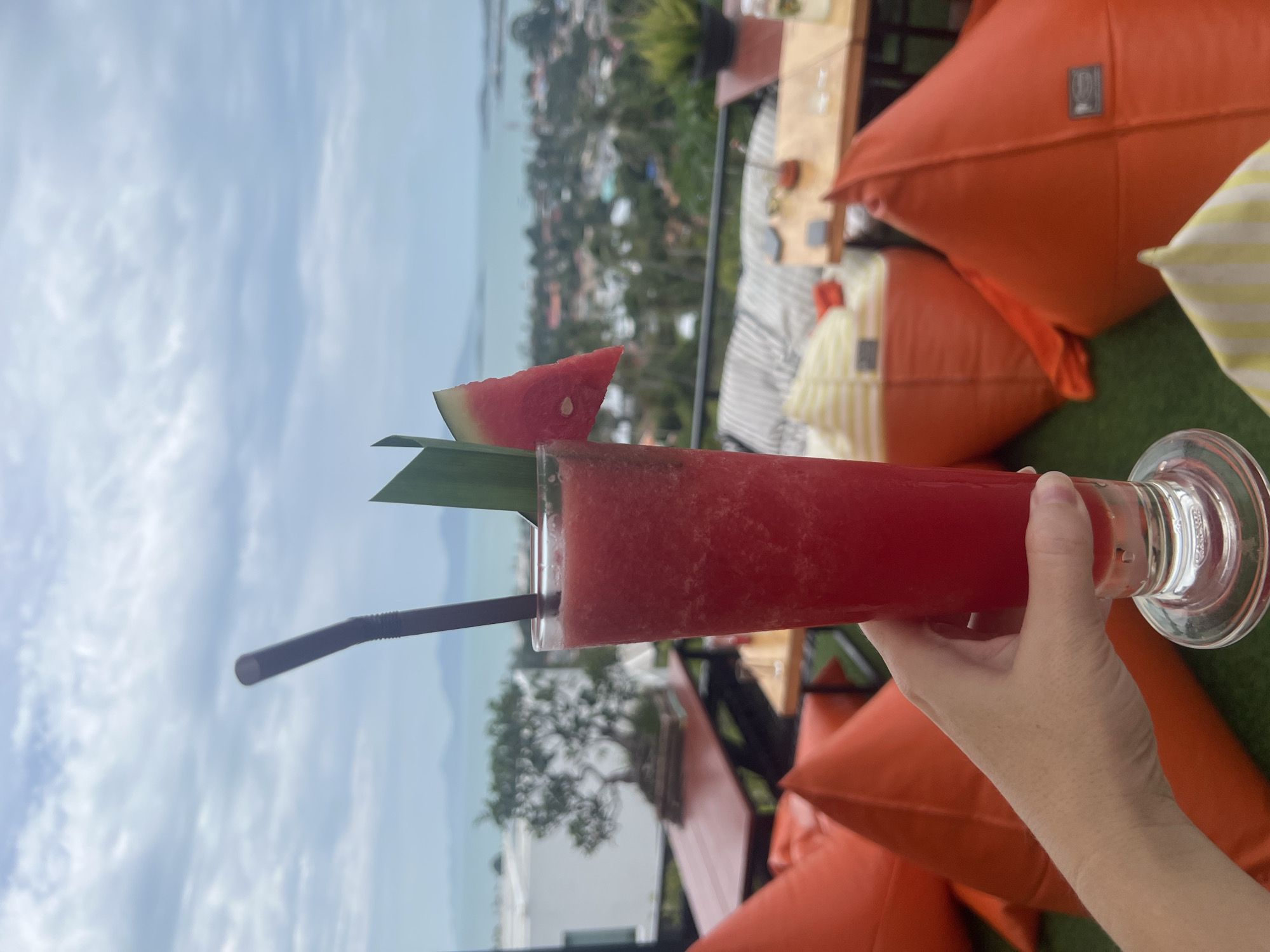 A hand holds a tall, clear glass filled with a vibrant red, blended beverage, likely a watermelon smoothie. A dark straw extends from the drink, and a slice of watermelon, along with a green leaf, is perched on the rim of the glass. The drink is presented against a scenic backdrop in Ko Samui District, Thailand.
In the foreground, several large, orange beanbag chairs are visible, suggesting a relaxed, outdoor seating area. Beyond these, wooden tables and chairs are set up, some with patrons or drinks. The middle ground reveals a panoramic view of a coastal town, characterized by houses nestled among lush green trees, leading to a body of water, likely the sea, under a cloudy sky. In the distance, faint hills or islands can be seen on the horizon. A small, meticulously pruned tree, possibly a bonsai, sits on a red table in the left portion of the image, adding an element of deliberate landscaping. The overall ambiance is that of a casual establishment offering scenic views, such as a cafe or restaurant with an outdoor terrace.