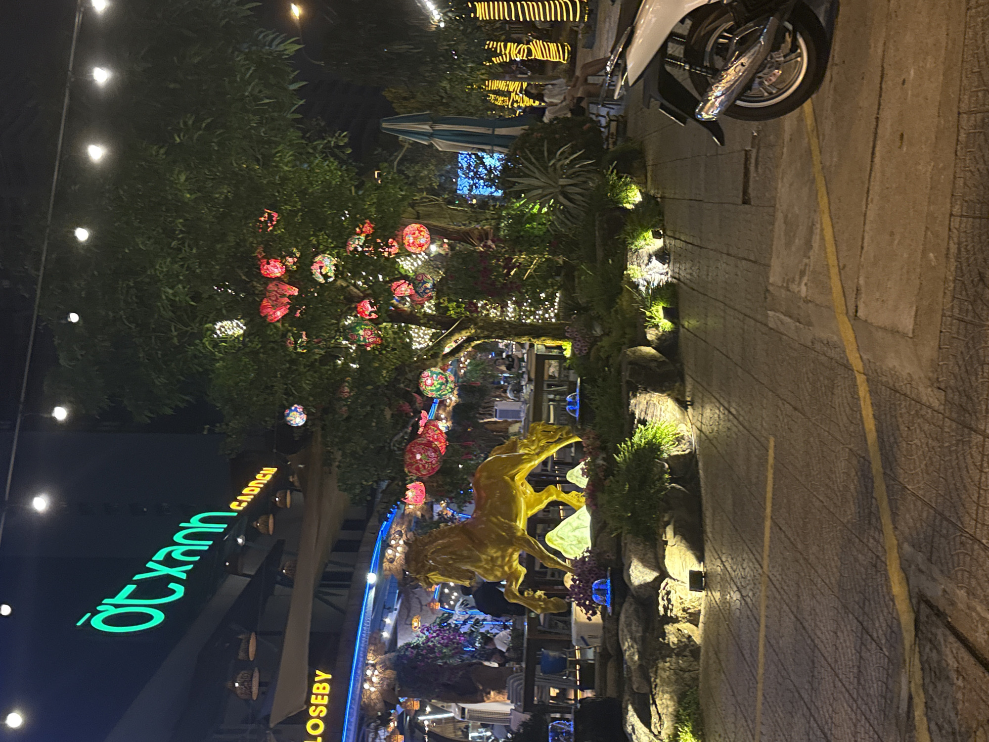 This nighttime image depicts a vibrant outdoor garden area, likely a restaurant or cafe, illuminated by numerous artificial lights. The setting is in Đà Nẵng, Vietnam, as indicated by visible text and general environment.
A central feature is a large tree adorned with many colorful lanterns, including red and multi-patterned designs, along with white string lights. Below this tree, a prominent golden statue of a horse stands dynamically, surrounded by lush green foliage and large rocks which are accentuated by small ground spotlights.
On the left, a building facade features a bright green neon sign displaying "ỚT XANH GARDEN", which translates to "Green Chili Garden". Below this, part of another sign, "LOSEBY", is visible. In the background, hints of an outdoor dining area can be seen, with people seated at tables and a blue and white striped umbrella.
A paved pathway or sidewalk with yellow lines runs along the right side of the scene, where the front wheel and part of a white scooter or motorcycle are parked. The overall ambiance is festive and inviting, with the dark night sky contrasting with the warm, abundant lighting. The image conveys a lively commercial establishment designed for evening enjoyment.