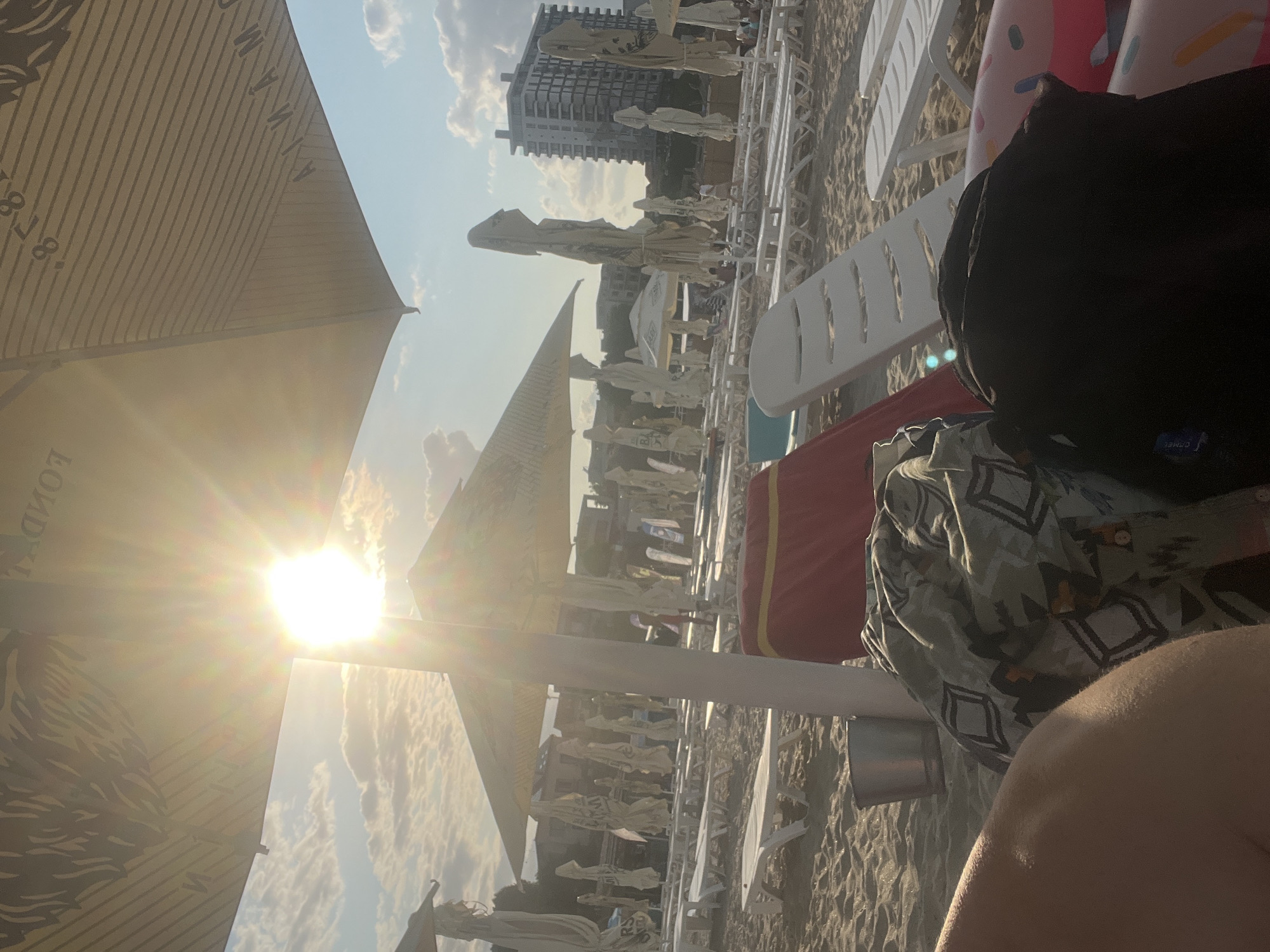 The media file depicts a beach scene in Constanța, Romania. The sun is shining brightly, suggesting it is daytime, likely mid-morning or afternoon, given the angle of the sun and the lighting. The sky is partly cloudy, with scattered white clouds visible.
The foreground is dominated by a beach umbrella, casting a shadow and partially obscuring the view. Below and to the right of the umbrella, there are beach chairs and umbrellas lined up on the sandy beach. One chair in the foreground has a person's leg resting on it, covered by a patterned garment. Another person's arm or shoulder is visible in the bottom right corner, with a hint of skin.
Behind the beach chairs, a tall, modern building with many windows is visible, suggesting the beach is located in a developed area, possibly near a resort or city. Other beach umbrellas are scattered further back, and some people can be seen in the distance, though they are not clearly distinguishable. The overall atmosphere appears to be relaxed and summery, characteristic of a beach vacation.
There is some visible text on the beach umbrella in the foreground, which appears to be a brand name or logo. Though partially obscured and blurred by the sun's glare, some letters can be deciphered, resembling "LONDON". Another umbrella further back seems to have text that is too blurry to read. The items visible on the beach chairs include a black backpack with a blue tag, a red bag with a yellow stripe, and a patterned blanket or towel. The scene conveys a sense of leisure and enjoyment of a sunny day at the beach.