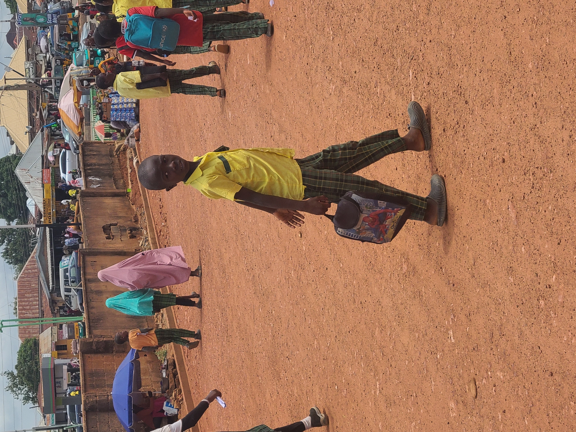 The image shows a group of school children in Jos, Nigeria, walking on a dirt road. They are wearing yellow shirts and plaid pants, which is likely their school uniform. One child is carrying a Spider-Man themed bag. The background shows a bustling urban scene with cars, shops, and other people. A woman in a pink hijab and another in a green hijab are also seen among the children. The road is unpaved, and the buildings in the background are typical of a developing urban area.