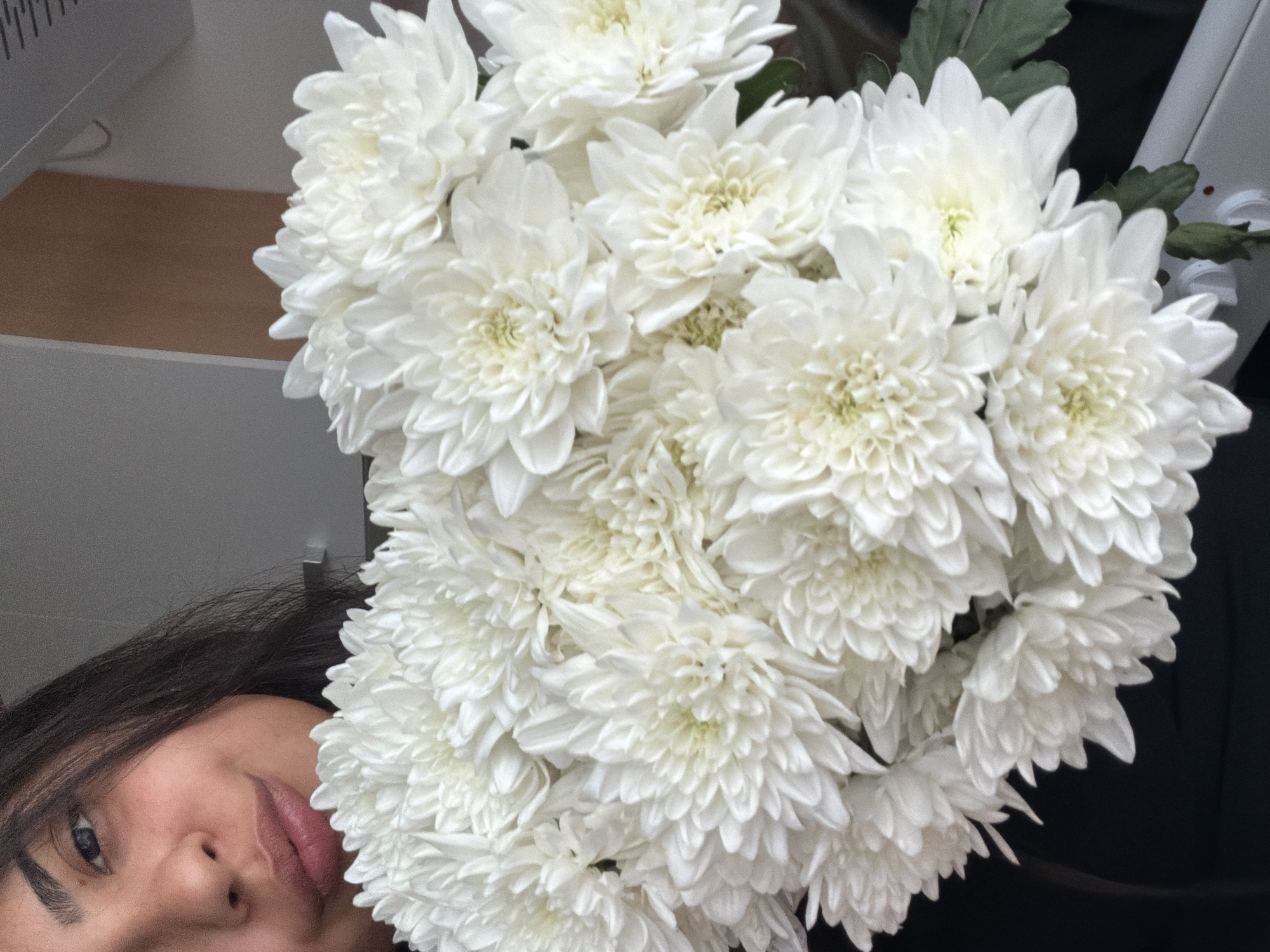 A close-up, medium shot captures a person partially visible, holding a large bouquet of white chrysanthemums. The person's face is positioned in the upper left quadrant of the frame, with their dark hair falling across their forehead and shoulder. They have dark eyebrows and a subtle smile. The chrysanthemums dominate the majority of the image, their densely packed petals creating a soft, textured mass of white. Some green leaves are visible amongst the flowers. The background is blurred and consists of neutral tones, suggesting an indoor setting. There is no discernible information about the location in Duisburg, Germany.