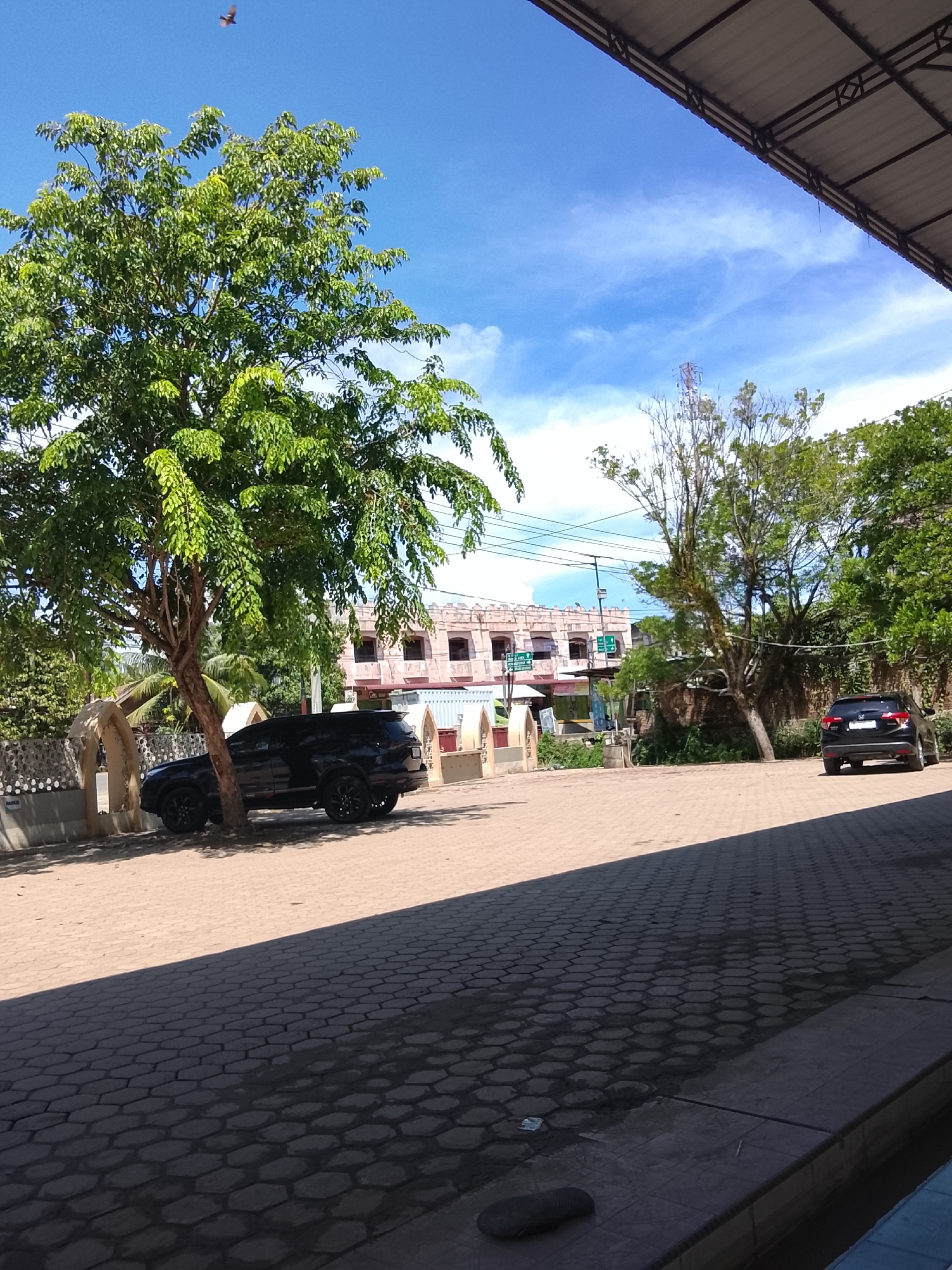 The image depicts a street scene in Meunasah Raya, Indonesia, on a clear, sunny day with a blue sky and scattered clouds. The foreground is dominated by a paved walkway with a hexagonal pattern, casting a large shadow. To the left, a large, leafy green tree stands prominently. Beside the tree, a black SUV is parked. In the background, a light pink building with multiple windows and a tiled roof is visible, suggesting it might be a residential or commercial structure. Further back, more trees and vegetation can be seen, along with power lines and a distant telecommunications tower. A small bird is captured in flight in the upper left portion of the sky. The overall atmosphere is one of a typical day in a tropical urban environment.