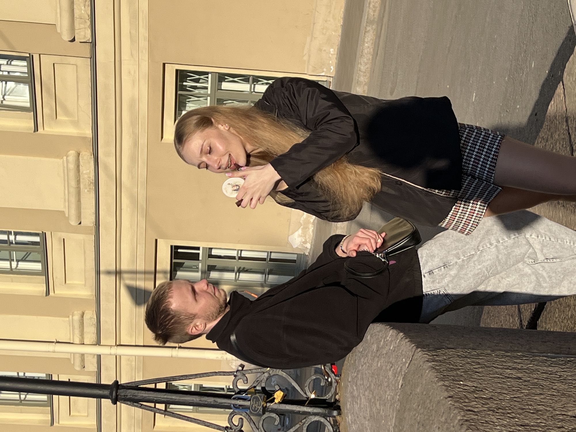 A street scene shows a blonde woman standing while holding a round object in her hands and looking at it. Standing opposite her is a man with fair hair and a beard, who is looking towards the woman. He is positioned close to a stone ledge or barrier, near a metal fence decorated with padlocks. The background includes a pale yellow building with a series of windows, suggesting an urban environment in daylight. The scene is in Saint Petersburg, Russia.