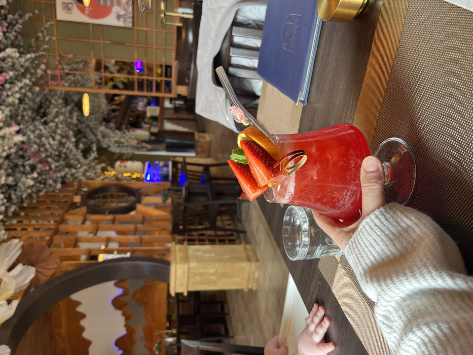 A person holds a red strawberry cocktail adorned with fruit garnishes in a stemmed glass. A clear glass and a baby's hand are visible beside the drink on a brown textured table. In the background, a restaurant interior features a decorative wall, plants, and seating. A blue menu is visible nearby. The scene has a blurred, artistic quality.