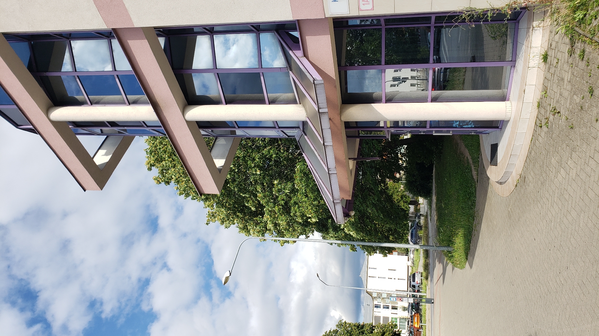 The image depicts the exterior of a modern building with a glass facade featuring purple frames, located in Prešov, Slovakia. The building is situated next to a sidewalk made of interlocking pavers and a grassy area with a mature tree. In the background, there are other buildings, parked cars, and streetlights, suggesting an urban or suburban environment. The sky is partially cloudy with visible white clouds against a blue background. The lighting indicates it is daytime, likely late morning or early afternoon, with the sun casting shadows. There are no people visible in the frame.