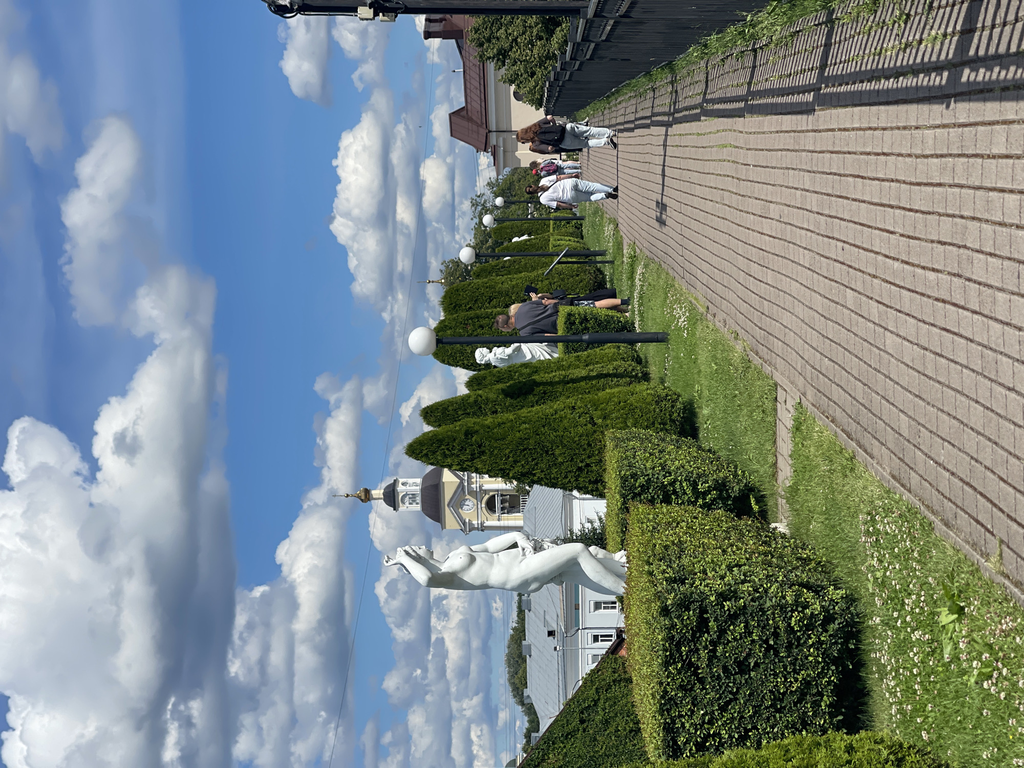 A paved pathway with a black metal fence on the right runs alongside a manicured garden on the left. The garden features a white classical statue of a nude figure, several tall evergreen trees, and neatly trimmed hedges. In the background, a yellow building with a clock tower, topped with a golden dome, is visible. Several people are walking along the pathway, dressed in casual clothing. The sky is blue with scattered white clouds. The scene appears to be in a park or a public garden. The general architecture and the presence of a Russian Orthodox church spire suggest a location in Russia. The architectural style and the overall aesthetic are consistent with the Vyborg Castle complex or its surrounding park areas in Vyborg, Russia.