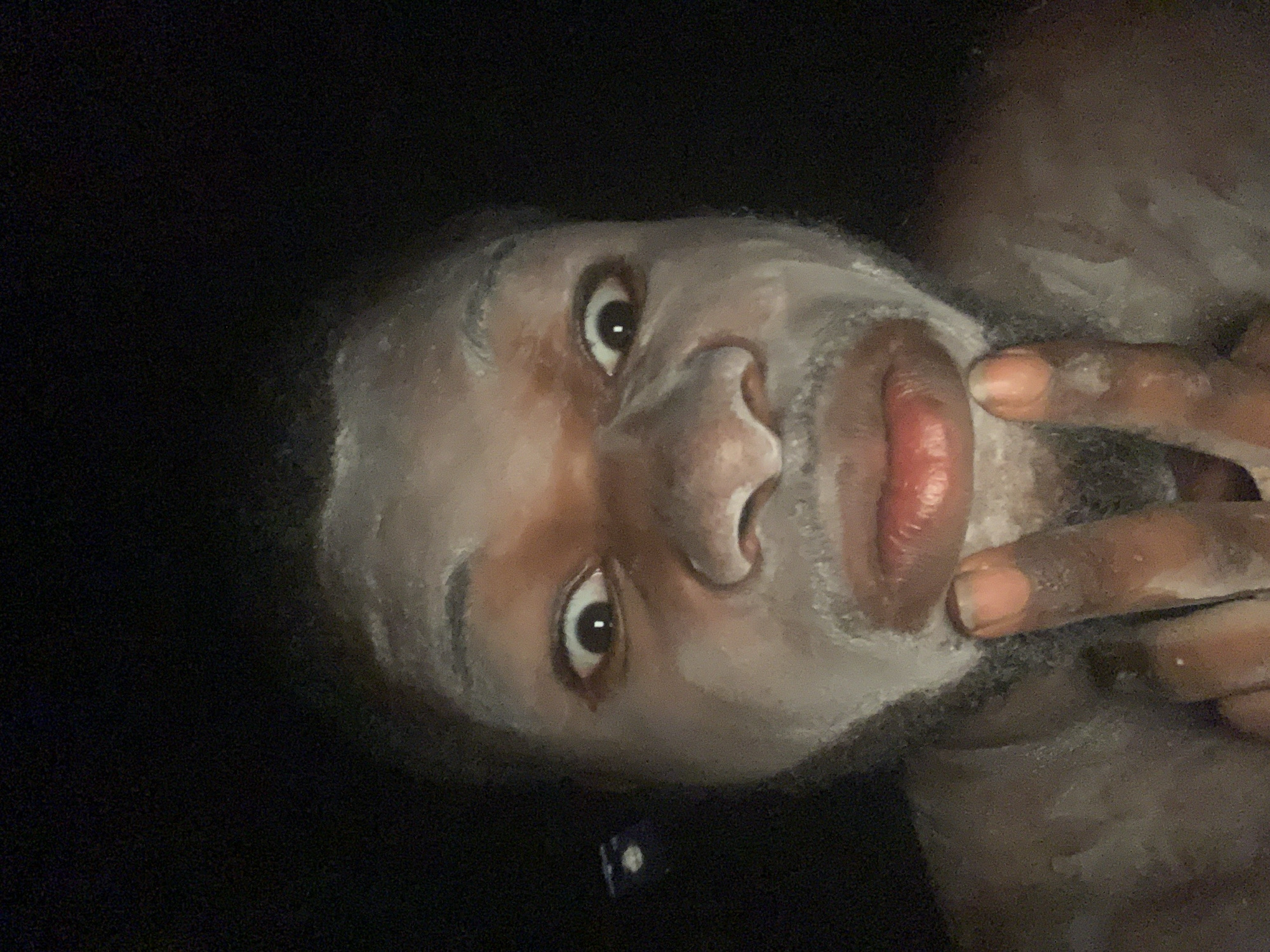 The media file presents a close-up portrait of an individual, likely an adult male with dark skin, in what appears to be an indoor setting in Obosi, Nigeria. The person's face, upper neck, and visible fingers are largely coated in a thick, white, foamy or pasty substance, indicating they are engaged in a personal hygiene activity such as washing their face or applying a face mask.
The subject's eyes are open wide, looking directly at the viewer with a neutral to slightly engaged expression. Their lips are visible, a rich reddish-brown, contrasting with the white substance. The surrounding background is predominantly dark and out of focus, suggesting low lighting conditions, possibly at night or in a dimly lit room. A small, blurry, dark rectangular object with faint white markings is faintly visible in the lower-left corner of the frame. No clear text is visible anywhere in the image. The image captures an intimate moment of self-care under subdued lighting.
