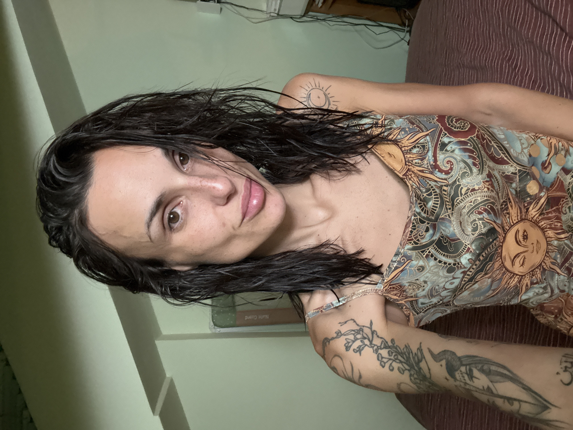 A woman with wet, dark hair and freckles is pictured from the chest up, looking at the camera. She is wearing a patterned sleeveless dress with sun and paisley designs. Her left arm, visible from the shoulder down, has several tattoos, including a branch with flowers, a figure with horns, and Arabic script. Her right shoulder also has a tattoo of a sun and crescent moon. The background is a room with light green walls, a white ceiling beam, and a bed with a striped maroon comforter. Electrical cords are visible on the right side of the frame. The image is a selfie taken indoors.