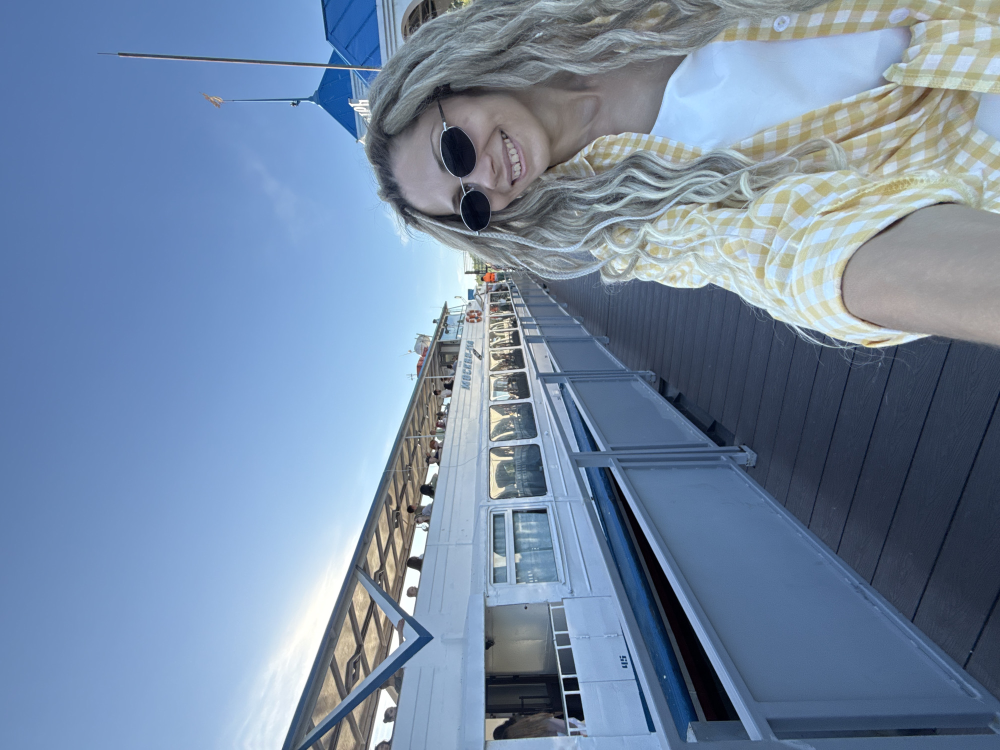 A detailed, forensic description of the scene in Dumino, Russia, shows a bright, clear day at a pier with a passenger boat. In the foreground, a young adult female with light skin and long, blonde dreadlocks/braids smiles, looking slightly past the camera to her right. She wears dark oval sunglasses with silver frames, a white top, and a yellow and white gingham plaid button-up shirt tied at the front. The shirt sleeves are rolled up.
To her left and in the midground, a white passenger riverboat named "МОСКВА-82" (MOSKVA-82/MOSCOW-82) is docked parallel to a dark grey composite or wooden pier. The boat features multiple rectangular windows along its lower deck and an open-air upper deck with a blue and white striped canopy supported by a metal framework. Several individuals are visible on the upper deck. The number "45" is displayed in blue on the boat's hull near the waterline. A light grey metal railing separates the pier from the boat.
In the far midground, behind the woman's head, a building with a distinctive blue roof is partially visible. A flagpole with a small, light-colored flag extends from the roof. The sky above is a clear, bright blue with minimal cloud cover, and sunlight suggests the sun is to the left of the frame.