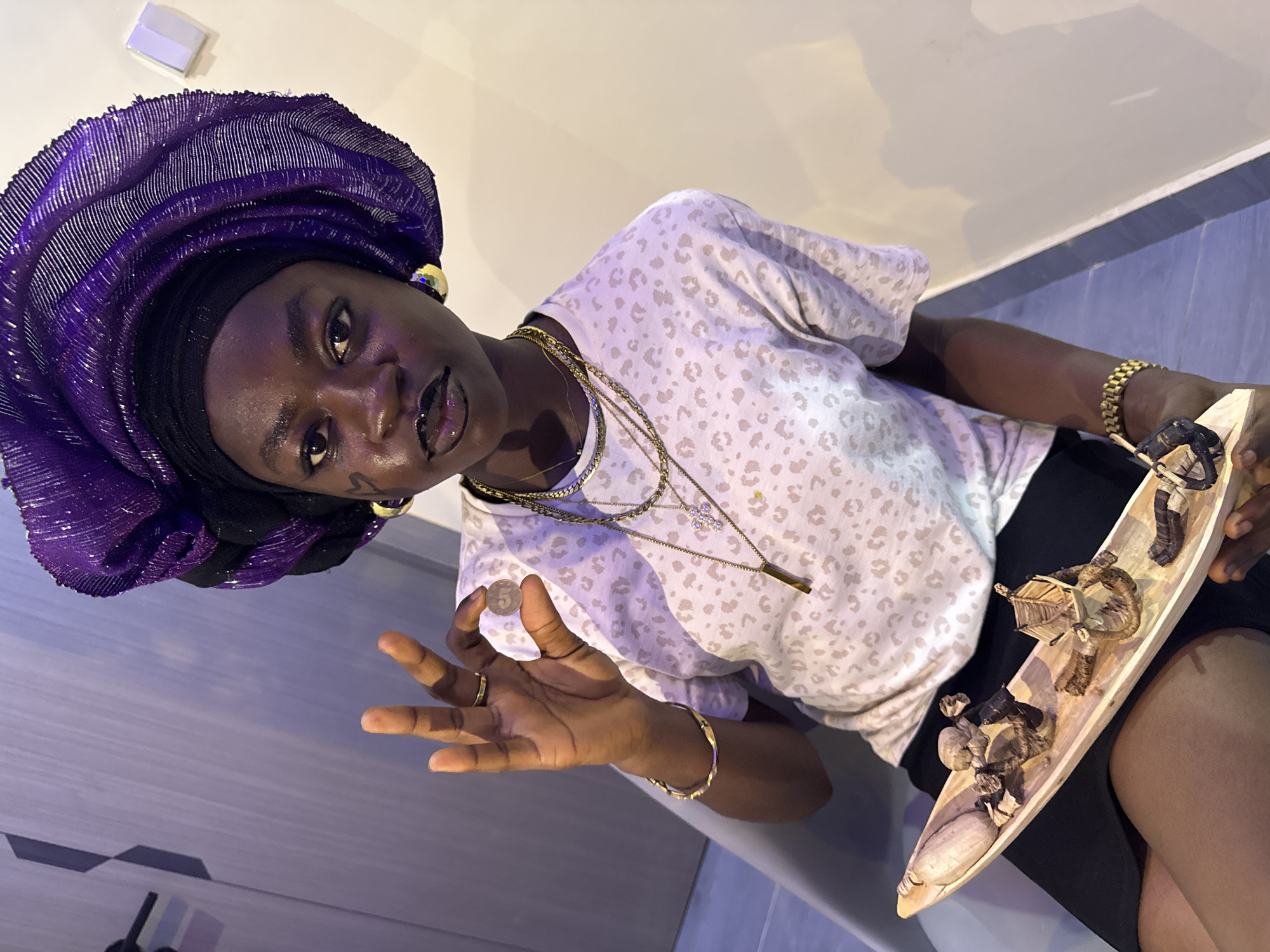 A person is pictured indoors. The person wears a purple headwrap, a white leopard print shirt, black shorts, and multiple gold necklaces and bracelets. The person holds a wooden tray with miniature sculptures. On the person's left cheek, there is a small drawing of a swirl. The person is holding up a five kobo Nigerian coin. The background is a light beige and blue wall. The location is Ajara, Nigeria.
