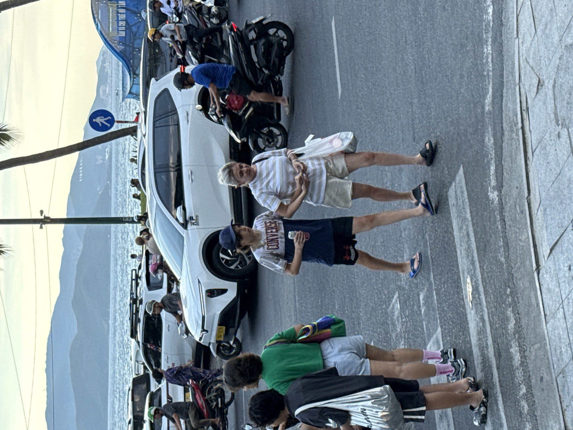 This media file captures a street scene in Nha Trang Ward, Vietnam, likely during daylight hours, possibly late afternoon given the light. The setting appears to be a busy street adjacent to a body of water, with mountains visible in the background. The atmosphere is lively and suggests an urban environment with significant pedestrian and vehicular traffic, predominantly motorbikes.
The primary subjects are several individuals, mostly young adults. They are engaged in various activities: some are standing and talking, while others are interacting with motorbikes. One person is leaning over a white car, possibly checking on it or retrieving something. Another individual is walking with a white bag.
Notable details include a pedestrian crossing sign with a blue circle and a white walking figure, indicating a designated crossing area. The ground is paved with concrete and asphalt. A large blue and white banner with text in a foreign language is visible in the background. Some individuals are wearing casual clothing, such as t-shirts and shorts, and several are wearing sandals or flip-flops. One person is wearing pink socks. The overall impression is that of a typical day in a bustling Vietnamese city, with people going about their business. The image is oriented vertically, suggesting it was taken with a phone in portrait mode.