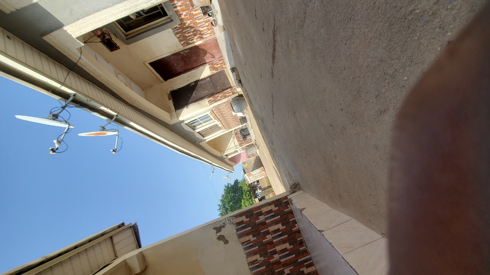 The image captures a street scene in Jalingo, Nigeria, likely in a residential or commercial area. In the foreground, a portion of a person's hand or arm is visible, obscuring the bottom part of the frame. The ground is a dirt or gravel surface, consistent with unpaved roads found in many parts of the city.
To the left, the side of a building is seen, featuring a tiled facade in shades of brown, orange, and white. This building has a light-colored roofline and appears to be part of a row of structures.
Dominating the center and right of the image is a row of connected buildings, likely shops or apartments. These buildings have pale-colored walls and cream-colored eaves. Multiple doorways are visible, some with dark-colored doors. The facades are decorated with a tiled pattern of brown, orange, and white above the doorways. Satellite dishes are mounted on the eaves of these buildings, indicating connectivity for television or internet services. One satellite dish is white and has a circular orange center. Another is entirely white.
In the background, a lush green tree stands tall, suggesting the presence of some vegetation in the area. A clear blue sky occupies the upper portion of the image. Scattered objects on the ground in front of the buildings include dark-colored plastic drums and what appear to be smaller containers or buckets. The overall impression is one of a typical urban environment in Jalingo, characterized by its construction styles and the presence of satellite technology.
