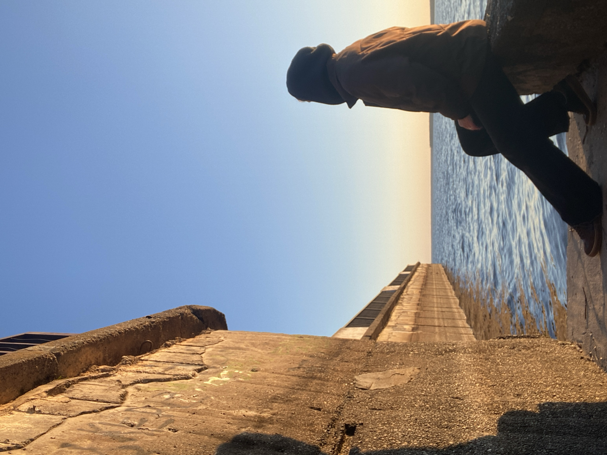 A man is leaning over a stone structure on the bank of the Kama River in Chaikovsky, Russia. The sky is clear and blue. The water of the river is rippled by the wind. The man is wearing a jacket, dark pants, and shoes. He is looking down at the water. The stone structure has stairs leading down to the water, and some of the stone slabs are cracked and discolored. The photo is taken from a low angle.