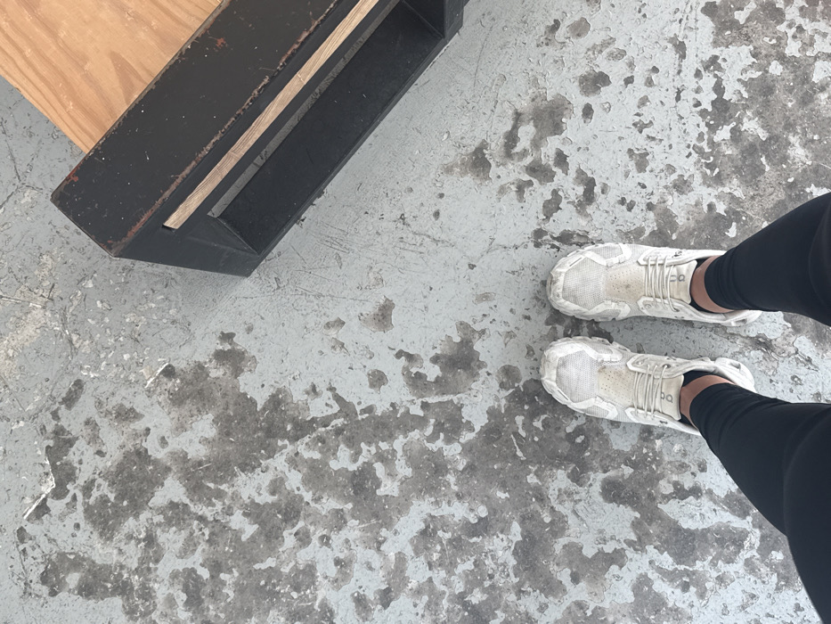 The image captures a high-angle view looking down at a person's legs and feet, standing on a distressed concrete floor in Puebla City, Mexico.
The primary subjects are the lower legs and feet of an individual, clad in black full-length athletic leggings and white athletic shoes. The shoes appear robust, with textured soles and uppers, and show signs of wear or dirt. On the outer side of both shoes, the brand logo "ON" is clearly visible.
The setting is characterized by a light grey concrete floor with numerous dark grey and black patches, suggesting worn paint, staining, or a naturally mottled, industrial-style surface. The texture of the floor is prominent, indicating an unpolished or heavily used area, possibly a gym, studio, or workshop.
In the upper left corner, a dark, rectangular object, likely a piece of furniture or equipment, is partially visible. It features a predominantly black surface, possibly painted wood or metal, with a lighter, natural wood insert or top portion. The object appears sturdy and functional.
The person is depicted in a standing posture, with no other discernible activity. The lighting is even, suggesting an indoor environment or an overcast day. No specific time of day or emotional cues are evident.