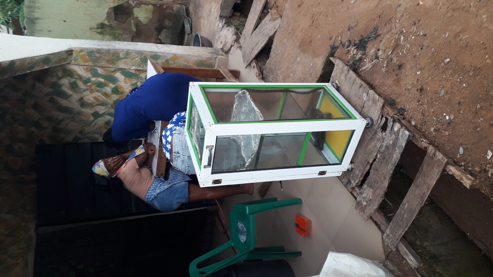 The image depicts an outdoor scene in Nkpor, Nigeria, near a building with a textured wall. In the foreground, a clear glass display case with a white and green frame stands on wooden planks over a drainage ditch. The case has two shelves, the top one lined with aluminum foil. A green plastic chair is visible to the left, with a small orange container and a plastic bag on it. Further left, a dark doorway suggests an entrance to a shop.
Two women are partially visible in the mid-ground, interacting near a wooden table. One woman, wearing a light brown shirt and a patterned headscarf, leans on the table, with her hand on her face. She is also wearing a blue and white checkered apron over denim shorts. The other woman, dressed in a bright blue dress, is bent over the table. A third person, partially visible from the back and wearing denim shorts and a blue and white patterned top, is also near the table. The ground is a mix of dirt and scattered debris. The overall impression is of a busy, informal commercial setting.