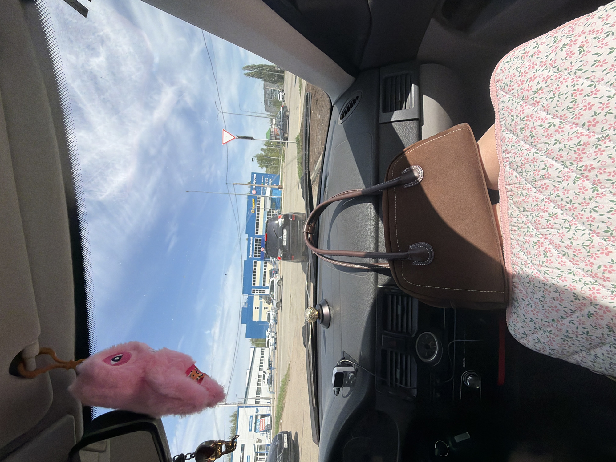 The view from inside a car shows a street scene in Chaikovsky, Russia. A triangular road sign is visible through the windshield. Buildings with blue and white exteriors are present on the right side of the road. A pink, plush bunny-shaped object hangs from the rearview mirror. A brown handbag rests on the dashboard, partially obscuring the center console. To the right of the handbag, a seat covered with a floral-patterned fabric is visible. Several vehicles are on the road, including at least two dark-colored cars.