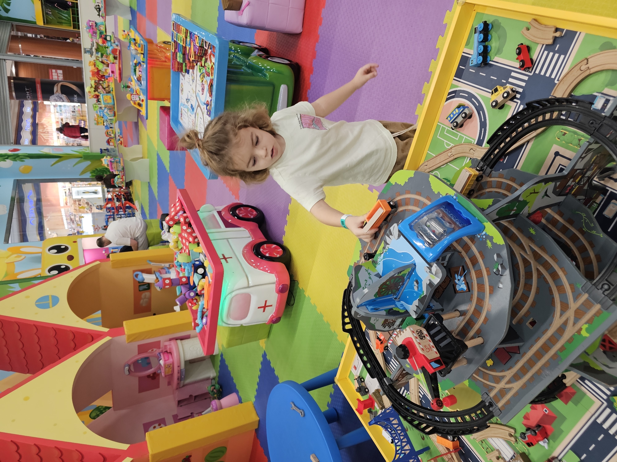 A child stands in a brightly colored indoor play area in Khimki, Russia. The child has light brown hair pulled back into a small bun, wears a light-colored t-shirt and beige pants, and has a teal wristband. The child is playing with a toy train and track set, holding an orange train car. The play area features a variety of soft play equipment, including a large playhouse, a toy ambulance, and a colorful foam floor mat with jigsaw-like edges. In the background, additional play structures and toys are visible. A person wearing a white shirt is visible farther back in the play area. The setting appears to be a commercial indoor play space.