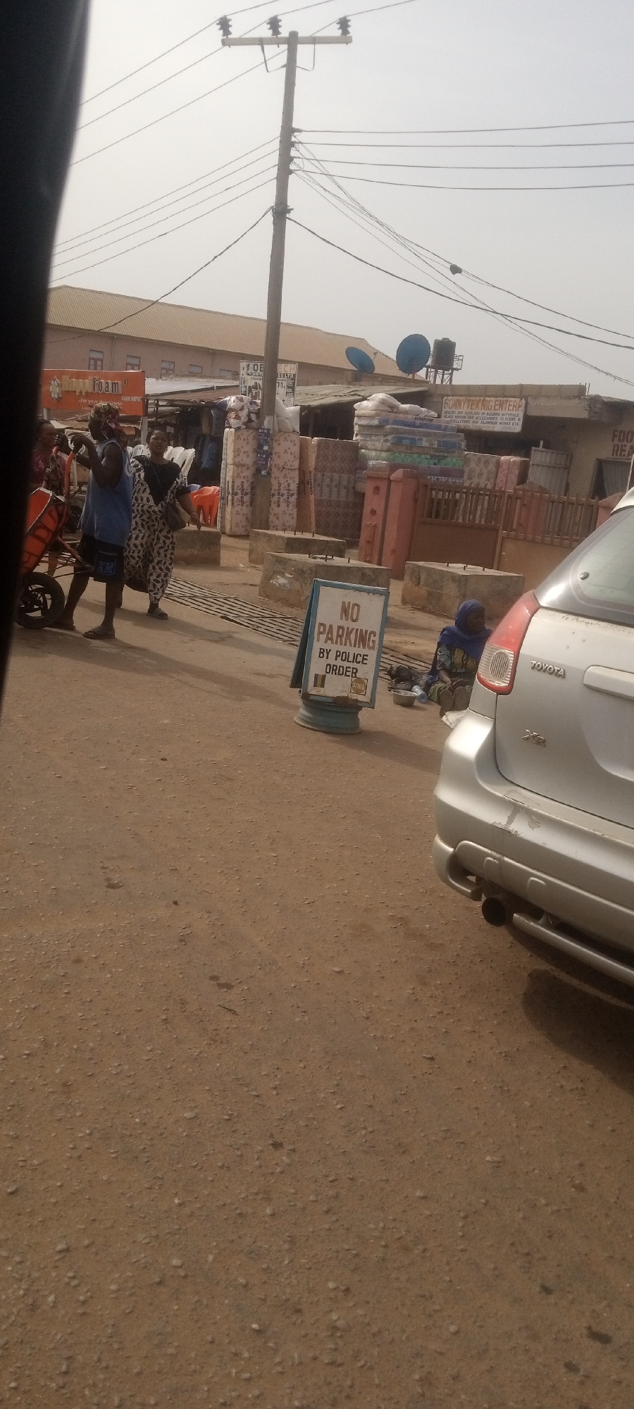 The image captures a vibrant street scene in Sabon Tasha, Nigeria, under a bright, somewhat hazy daytime sky. The immediate foreground shows a dirt and gravel road.
Several individuals are present, indicating daily activity. On the left, a man wearing a blue shirt and dark shorts is seen pushing a red, wheelbarrow-like cart. Beside him, a woman in a patterned, light-colored dress walks, looking towards the viewer. Further to the right and closer to the ground, a person wearing a blue head covering and colorful attire sits next to a silver Toyota XRS car, with a small bowl in front of them.
A white sign with a blue frame stands prominently in the middle of the road, clearly stating "NO PARKING BY POLICE ORDER," with a smaller "SWMO" logo below. The rear portion of the silver Toyota XRS car is visible on the right, facing away, showing some minor scuffs on the bumper.
The background features a bustling commercial area. Numerous mattresses or large bales of goods are stacked high outside various shops. Concrete barriers are placed along the roadside. Several signs are visible: a yellow banner for "HAPPY FOAM," a larger sign for "RONNYTEX ENTERPRISE" advertising "DEALERS AND SUPPLIERS OF BUILDING MATERIALS, PLASTICS, SANITARY WARES, GLAZIERS & INSTALLATORS AND ALUMINUM WORKS," and another partial sign reading "FOOD REAL." Overhead, a tall utility pole supports numerous power lines against the light sky. Two blue satellite dishes and a black water tank are mounted on the rooftops of the buildings. The scene conveys an impression of everyday commerce and street life in a busy setting.