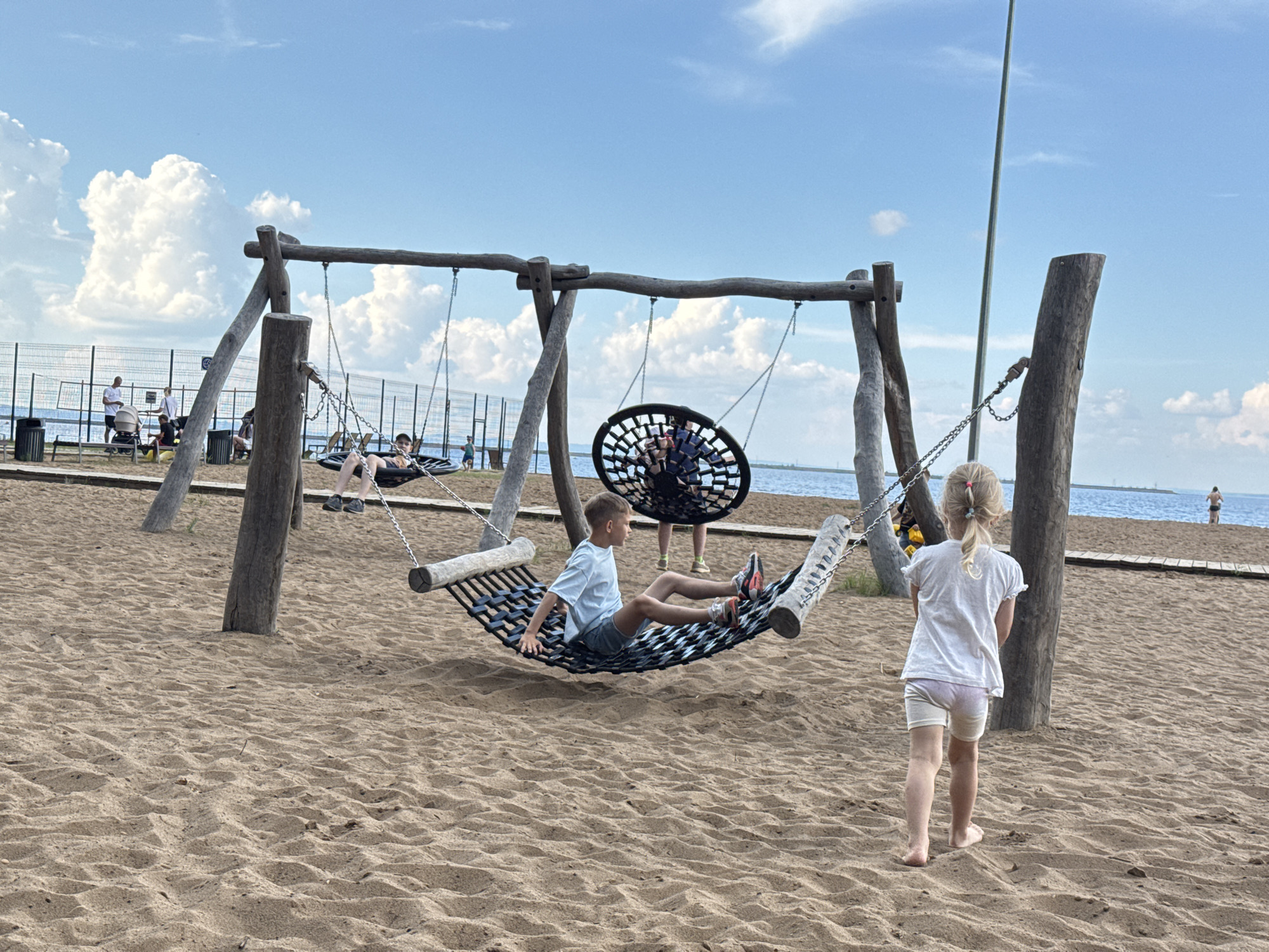 The image shows a sandy beach area in Chaikovsky, Russia, with a playground structure. The playground features a large wooden frame with several swings, including two suspended hammocks and one round net swing. In the foreground, a young boy in a light blue t-shirt and grey shorts is relaxing in one of the hammock swings. To the right, a young girl with blonde hair in a ponytail, wearing a white t-shirt and light-colored shorts, walks away from the viewer. In the background, there is a body of water, likely the Kama River, with a fence and some people near it. The sky is blue with scattered white clouds. The overall scene depicts a recreational area on a sunny day.