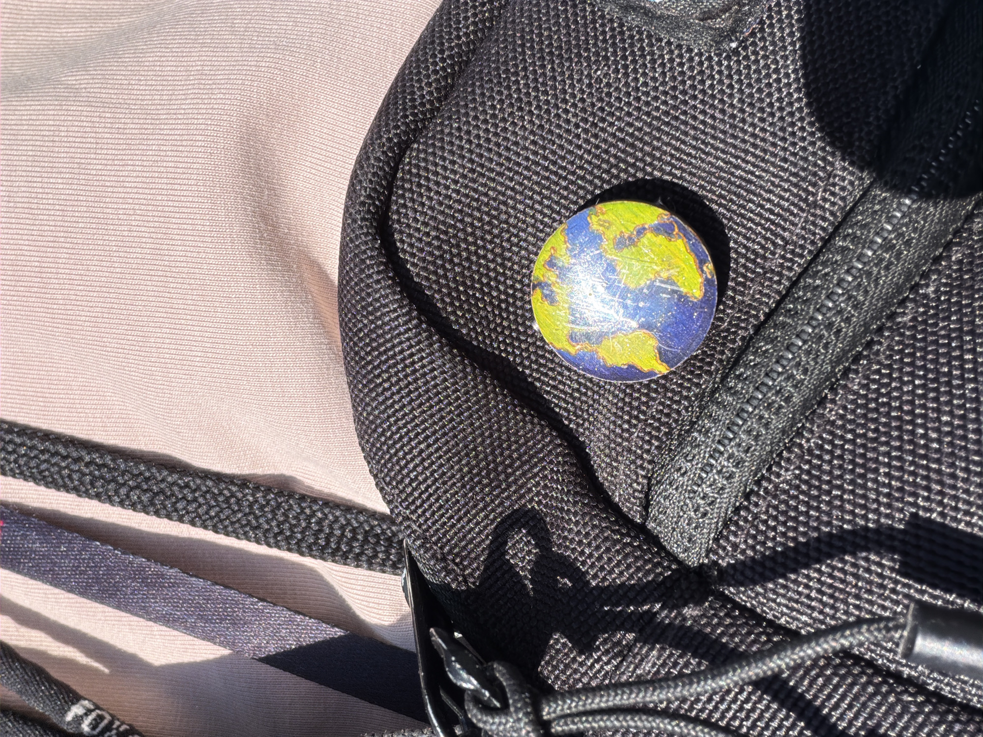 The image shows a close-up of a black backpack. Affixed to the backpack is a small, round object resembling a stylized globe, with green areas representing landmasses and blue areas representing water. The backpack is partially situated on a light tan surface. The presence of a "FOX" logo on a nearby strap suggests it might be a product of Fox Racing. The location of the image is unknown, and there is no indication of it being in Oreshniki, Russia.