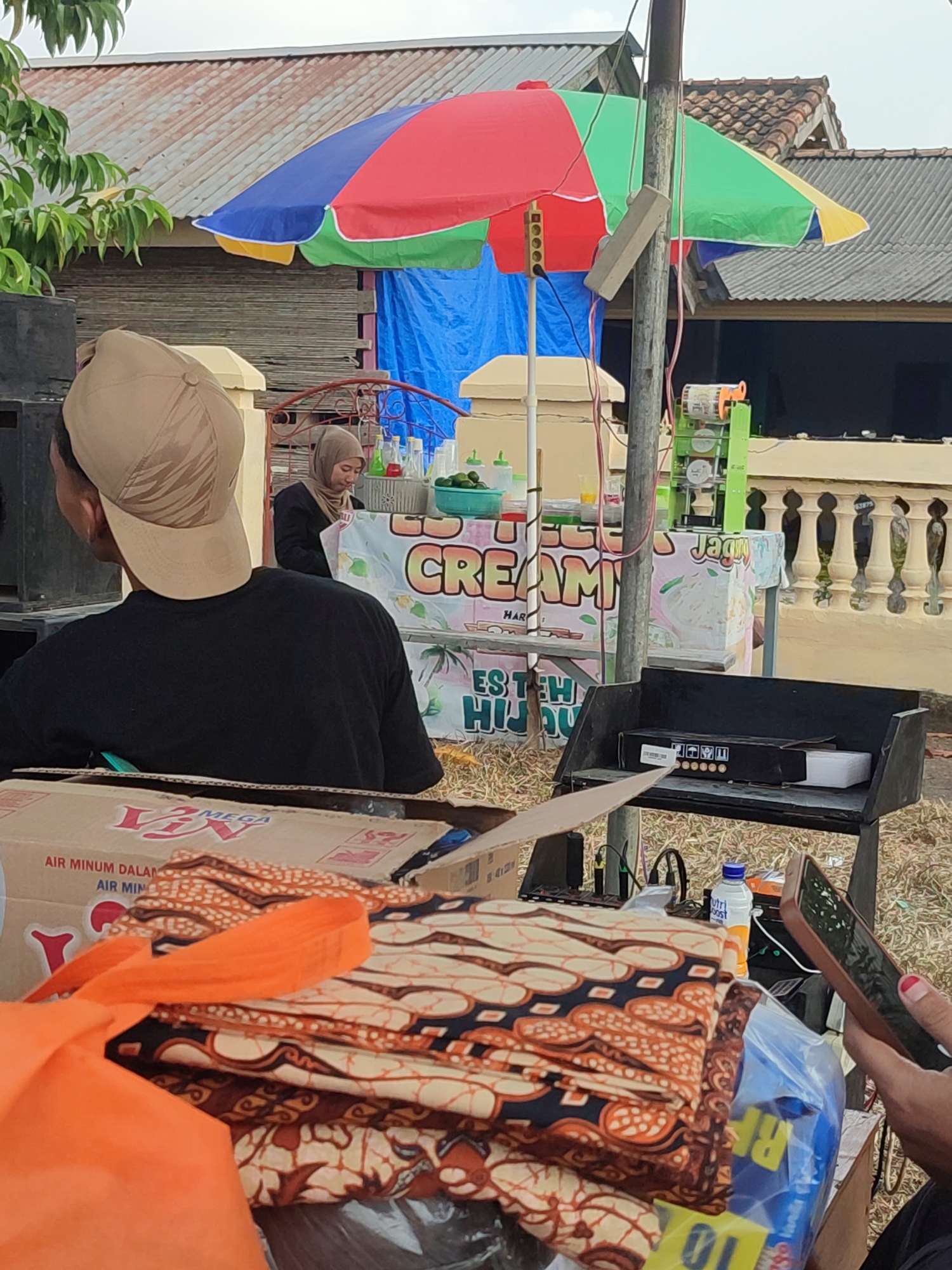 The image depicts an outdoor scene in Nusa Jaya, Indonesia, likely a street market or a temporary stall. In the foreground, a person wearing a black t-shirt and a tan baseball cap is viewed from behind, facing away from the camera. They are positioned near a large black speaker. In front of them, a stack of folded batik fabric with orange and dark brown patterns is visible, along with an orange tote bag. To the right, a person's hand holding a smartphone is visible, with a partial view of their arm and nail polish.
Behind the person in the cap, a small food stall is set up. A colorful umbrella, with red, blue, yellow, and green panels, provides shade over the stall. A woman wearing a light brown hijab and a black top is working at the stall, with various bottles and containers of liquids and ingredients displayed. Signs are visible on the stall, with text including "CREAMY" and "ES TEH HIJAU". To the right of the stall, a green juicing machine is present. The background shows a building with a corrugated metal roof and a wall. The ground is covered with dry grass and dirt. A shelf unit holds various items, including a black box and what appears to be electronic equipment. A bottle of "NutriBoost" is also visible. The overall impression is a bustling, informal commercial setting.