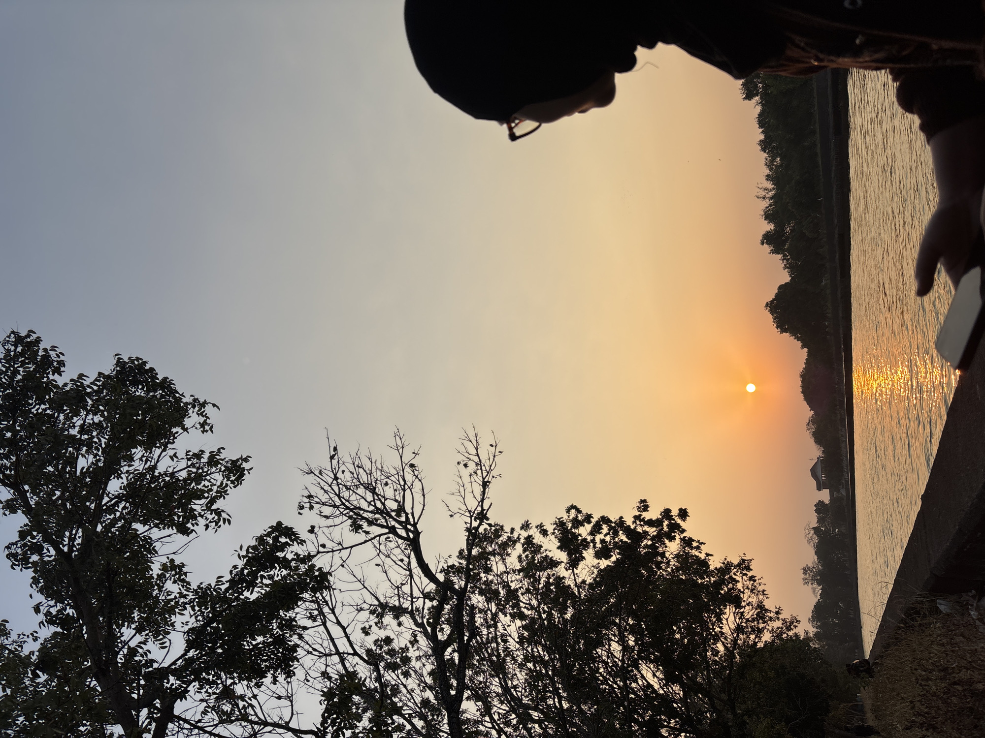 A person wearing a hijab and glasses is seen in profile, looking out at a sunset over a body of water. The sun, a bright orange orb, is low on the horizon, casting a warm glow across the sky and reflecting on the water's surface. Silhouetted trees line the far bank of the water, with a building visible in the distance. The foreground shows a wall bordering the water, with dry grass and some discarded items on the bank. The image appears to be taken in a park or natural setting, possibly in the city of Panyingkiran, Indonesia, given the atmospheric conditions and vegetation. The overall mood is peaceful and serene.