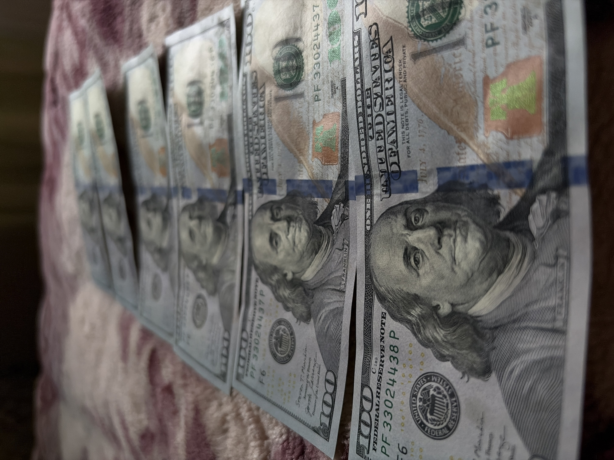 A close-up, low-angle shot shows a fan of United States one hundred dollar bills. The bills are stacked in descending order, with the top bill slightly out of focus. The bills are laid out on a textured, rose-colored surface, possibly a blanket or rug. The bills are oriented horizontally with the portraits of Benjamin Franklin facing upwards. The lighting is dim, creating shadows and highlighting the intricate details of the currency.