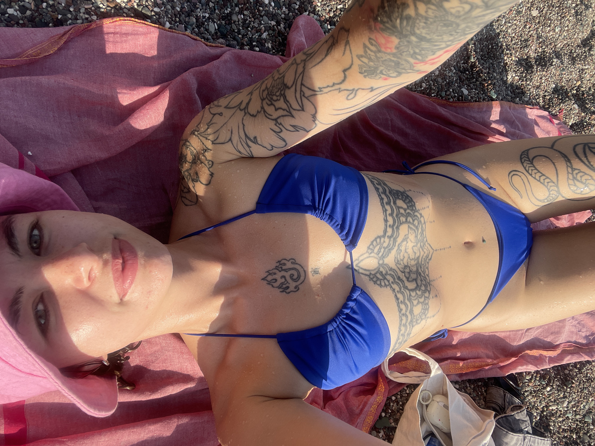 A woman is reclining on a reddish pink blanket on a pebble beach. She is wearing a bright blue bikini and a pink sun hat. She has multiple tattoos on her arms, chest, and abdomen. Her left arm is raised, possibly taking a selfie. The background shows small pebbles and what appears to be a bag. Based on the context provided, this location is in Rafailovići, Montenegro.
