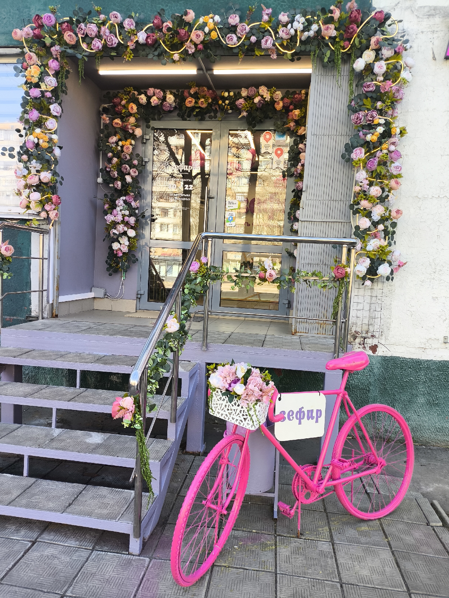 The image shows a retail location in Khimki, Russia. The storefront is decorated with an archway of artificial flowers above the entrance and along the edges. The entrance has double glass doors. A pink bicycle with a basket of flowers is parked in front of the entrance, and it has a sign on the bike with the word "зефир" (Zefir) in Russian written on it. The steps leading up to the entrance are painted a pale purple. The walls on either side of the doorway are white and gray.