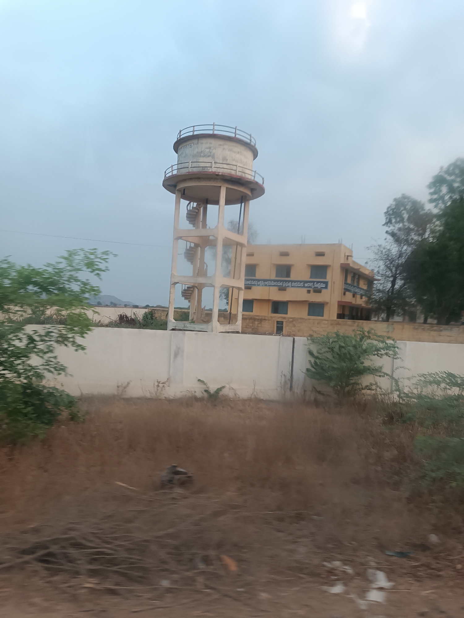 The image depicts a static scene in Kottalapalli, India, characterized by a prominent concrete water tower and a multi-story building under an overcast sky. In the foreground, dry, sparse vegetation and brown earth with scattered debris cover the ground. A light-colored concrete wall spans horizontally across the mid-ground. Behind this wall, a tall, light-beige concrete water tower with a cylindrical tank at its apex and an exterior spiral staircase stands centrally. To the right of the water tower, a two-to-three-story building with a light yellow facade and blue-framed windows is visible. Signs or banners with text in a local script are affixed to the building's upper level. Taller trees with dark green foliage are present to the right of the building. In the distant left background, faint outlines of hills are visible. No individuals or active interactions are present in the scene. The sky is uniformly light grey, with a slightly brighter patch in the upper right quadrant.